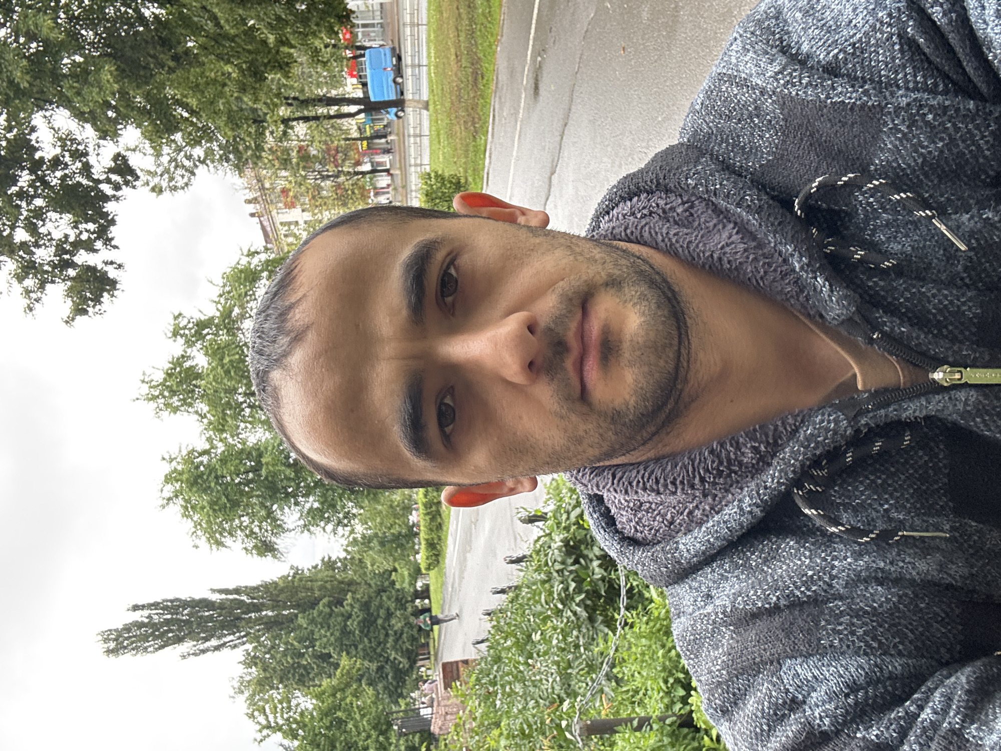 The image shows a man taking a selfie outdoors in what appears to be a city environment. He is wearing a grey and black patterned hooded jacket over a tan shirt. The background suggests a street or park area with trees, a paved pathway, and buildings visible in the distance. There are other people and a blue van in the background. The weather appears overcast. The specific location is identified as Ufa, Russia, though no specific landmarks are identifiable to confirm this.