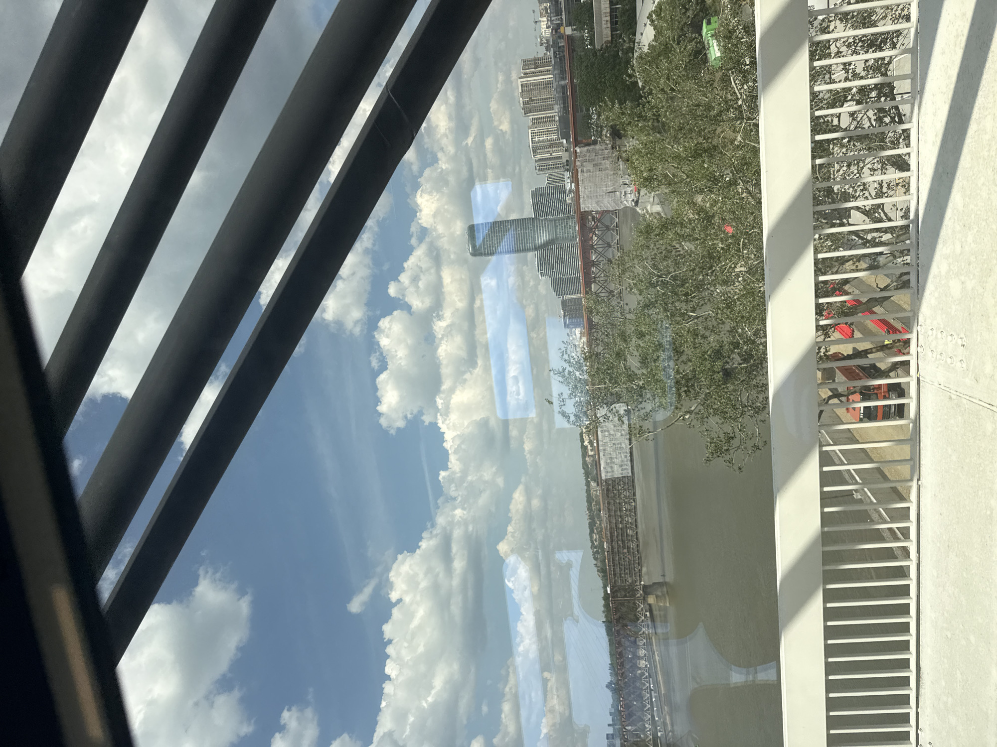 The image appears to be taken from inside a building looking out through a window. Slatted metal bars are visible on the left side of the frame, and a white metal railing is on the right side. The window's reflection shows a city skyline with a river, and the cloudy sky is reflected in the water. Buildings are visible in the skyline along with trees, and the bridge construction appears to be the Branko's Bridge. This suggests a location in Belgrade, Serbia, likely overlooking the Sava River.