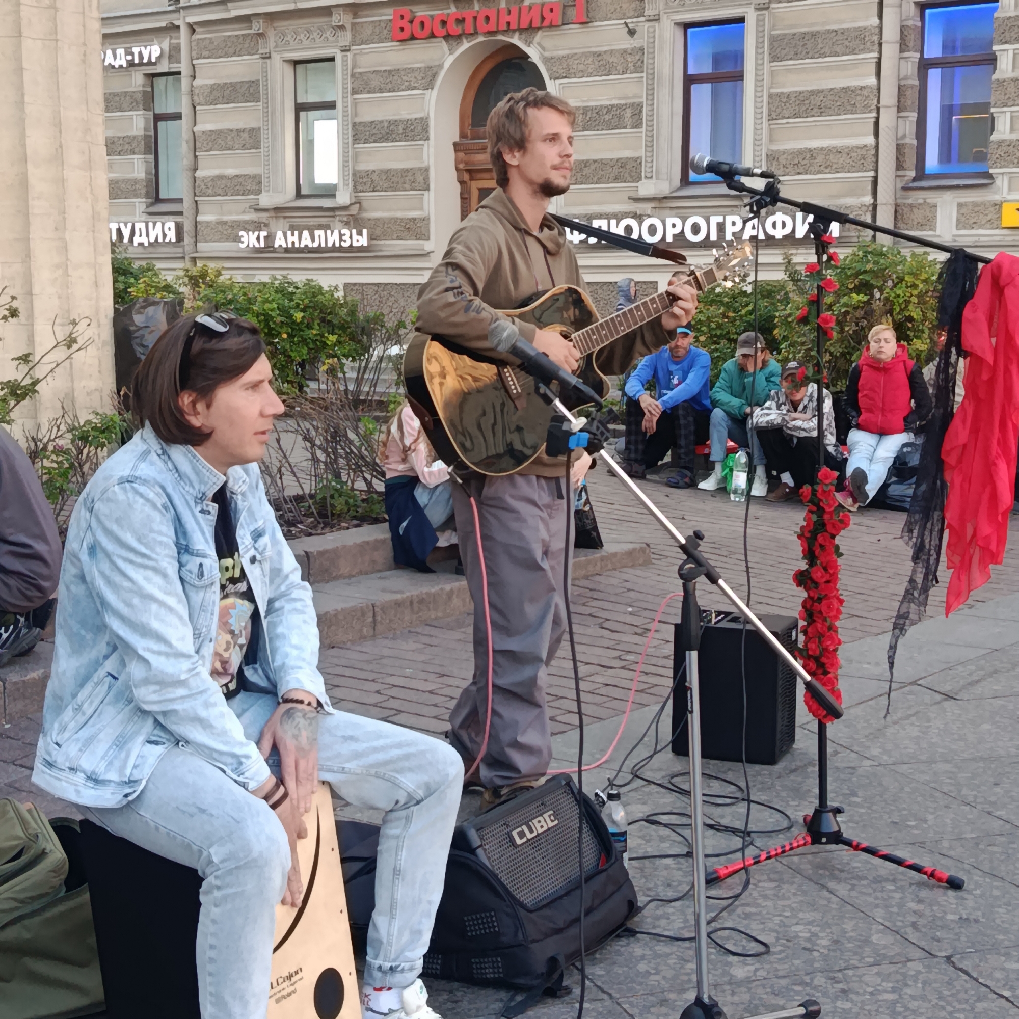 Two men are performing music on a city street, likely in Saint Petersburg, Russia, given the Cyrillic signage visible on the buildings in the background. The building facade features elaborate architectural details and windows with blue and white interiors. One man, positioned in the foreground on the left, is seated and playing a cajón drum. He is wearing a light blue denim jacket, a black t-shirt with a graphic print, and light blue jeans. He has dark hair and sunglasses perched on his head. The other man, standing behind him and to the right, is playing an acoustic guitar and singing into a microphone. He is wearing a brown hooded sweatshirt and grey trousers.
Several other individuals are seated on steps in the background, appearing to be an audience. They are dressed in casual clothing. To the right of the performers, a microphone stand is decorated with a string of red artificial roses and black lace. A black amplifier sits on the ground near the microphone stand. The ground is paved with grey cobblestones. The overall scene depicts a street performance taking place outdoors on a paved urban area with buildings in the background.