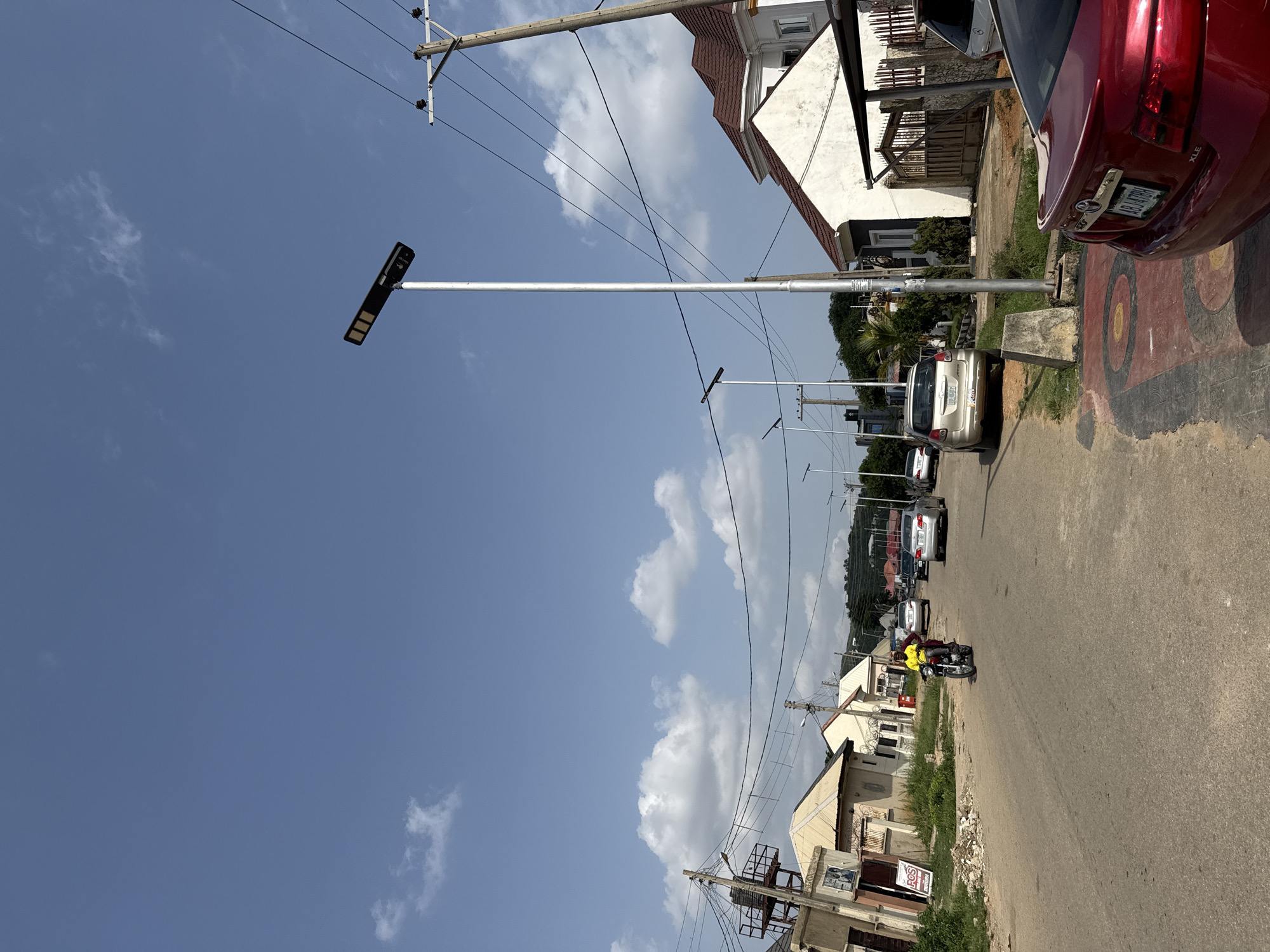 The media file depicts a sunny day in a residential area in Abuja, Nigeria. The scene is oriented vertically, showcasing a street lined with buildings and power lines. Dominating the foreground is a tall pole with a solar-powered street lamp. Further down the road, several cars are parked and in motion. A motorcycle with two occupants, one wearing a bright yellow shirt, is traveling down the street.
The sky is a clear blue with scattered white clouds, indicating fair weather. The architecture of the buildings suggests a tropical or subtropical climate. Power lines are a prominent feature, running across the frame, supported by wooden utility poles.
Visible text on a red car license plate reads "ABJ 47FH". A sign on a building also has text, partially obscured but appears to say "FOS". The overall atmosphere is that of a typical day in a suburban or urban neighborhood. The lighting suggests it is daytime, likely mid-morning or afternoon.
