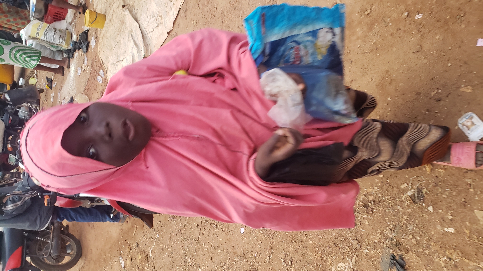 A young girl wearing a pink hijab and patterned skirt stands in an outdoor market, possibly in Kakuri Gwari, Nigeria. She is holding a blue plastic bag in her right hand, which appears to contain some items. Her left hand is also visible, holding a white plastic bag. She is wearing pink sandals. In the background, there are motorcycles and other market activities. The ground is covered with dirt and some scattered debris. The overall lighting suggests it is daytime.