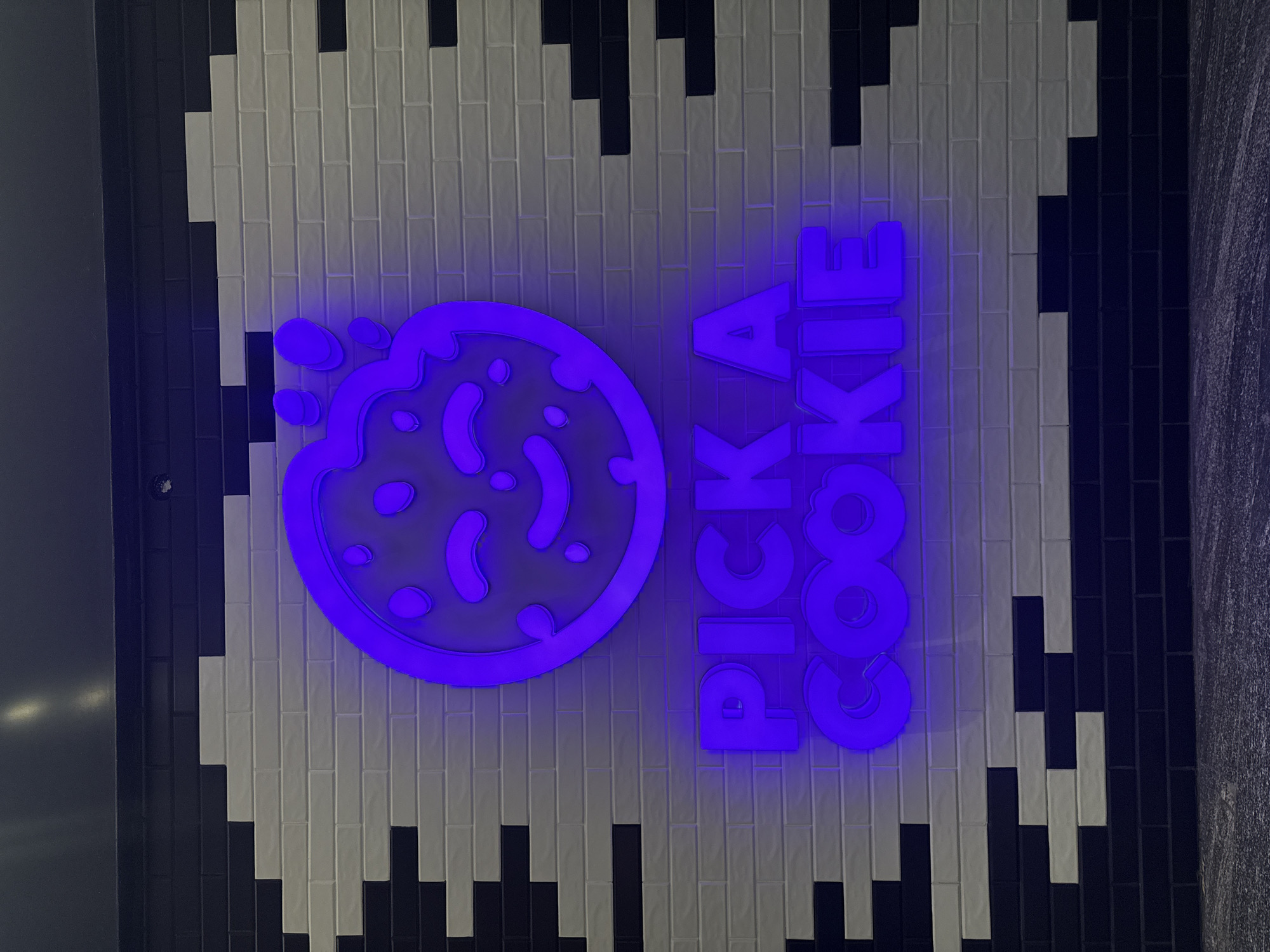 A vertical, close-up shot depicts a "PICK A COOKIE" illuminated sign on a textured wall. The sign features a cookie graphic to the left and the text "PICK A COOKIE" in a stacked arrangement to the right, all lit in a vibrant blue. The wall behind the sign is patterned with vertically aligned, alternating black and white tiles. The overall aesthetic suggests a modern or playful branding for a cookie business. The location is Puebla, Mexico.