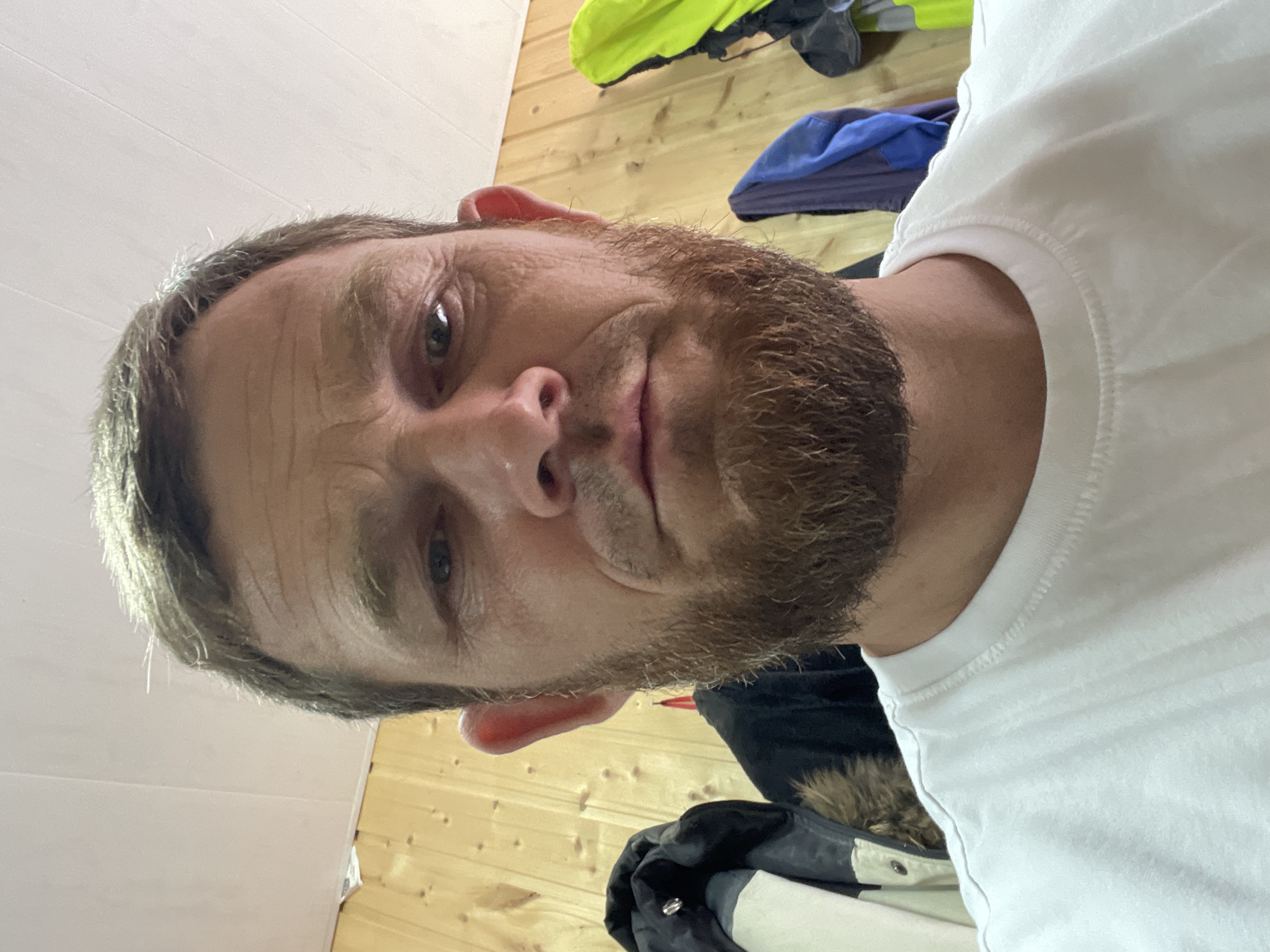 The image is a close-up selfie of a man with fair skin, short brown hair, and a light brown beard. He is wearing a white t-shirt. The background consists of wooden paneling on the walls and a white ceiling. Several articles of clothing, including jackets and what appears to be a hi-vis vest, are hanging on hooks on the wall behind him. The image was taken indoors. There is no indication of the location being in the city of Krasnodar, Russia.