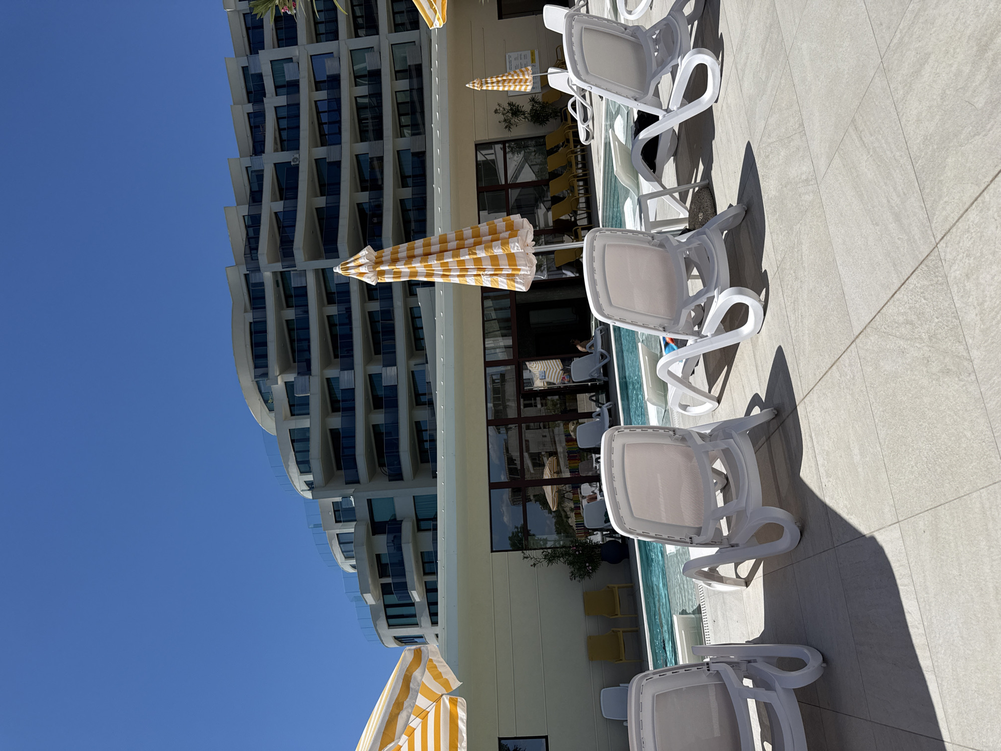 The image shows a resort swimming pool area on a sunny day. In the foreground, several white plastic lounge chairs are arranged on a light-colored tiled patio. To the left, part of a yellow and white striped umbrella is visible. Beyond the lounge chairs and a portion of the pool, a long, cream-colored building with multiple levels and balconies stretches across the frame. The building has a modern, curved architectural design. In the background, a clear blue sky dominates the upper portion of the image. The pool itself is visible with clear blue water. The overall scene depicts a tranquil, resort-like setting with amenities for relaxation and leisure.