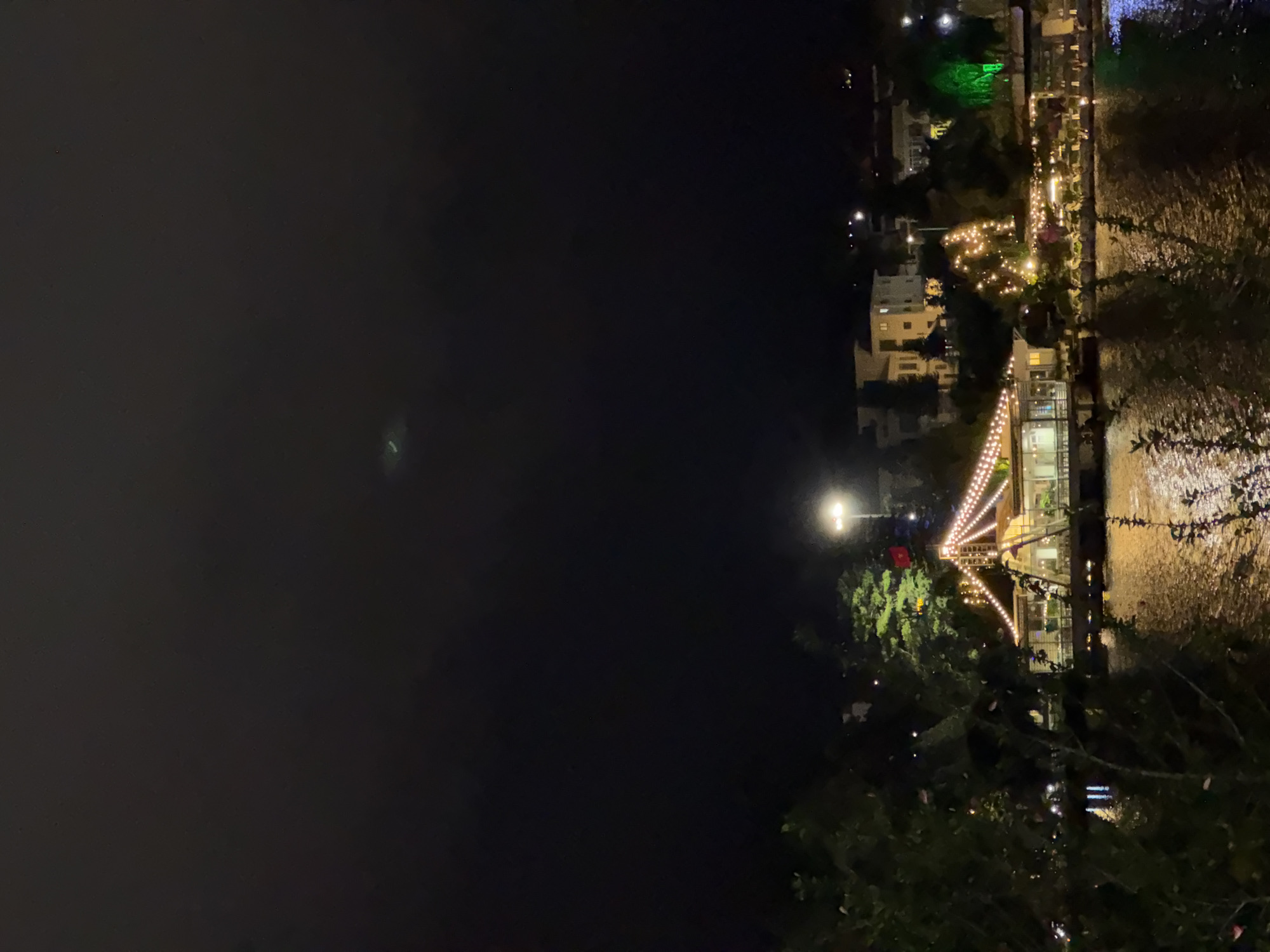 The image depicts a night scene in Da Lat, Vietnam, likely along Xuan Huong Lake. A brightly lit building, possibly a restaurant or event venue, sits on the edge of a body of water, its lights reflecting on the rippling surface. The building is adorned with numerous string lights, creating a festive atmosphere. In the background, residential buildings and trees are visible, some illuminated by streetlights. The sky is dark and cloudy, with a faint light streak possibly indicating an airplane or distant cloud formation. Lush foliage frames the left side of the image.
