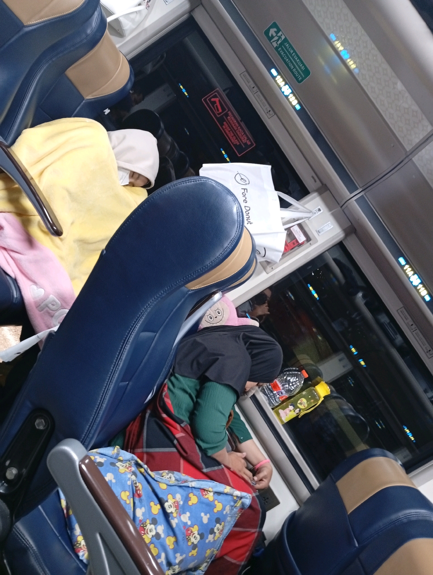 The image shows the interior of a train, likely on a journey. The perspective is from a seated position, looking forward and slightly upwards. In the foreground, a dark blue and beige train seat is prominent, with part of another identical seat to the left. A person is partially visible in the yellow blanket, covered by a light-colored hood. To the left of the yellow blanket, a pink blanket with a heart and lettering is visible.
Further into the scene, on the right, another person is seated, wearing a black headscarf and a green long-sleeved shirt. This person is leaning forward, with their left arm resting on their lap, and a red and black plaid blanket is draped over their legs. A plastic water bottle with a yellow cap and a partially filled water bottle with a red cap are on a surface to their right.
Above the seats, various signs and displays are visible. A green sign with white text reads "JALUR EVAKUASI" and "EVACUATION ROUTE" with an arrow pointing left. Below this, a digital display shows numbers and letters in blue and yellow. Another red sign with white text is also present.
The windows of the train show a dark, reflective exterior, with some lights visible, suggesting it is nighttime or the train is in a tunnel. The overall environment suggests a mode of transportation with multiple passengers.
Given that Waruduwur is a village in Brebes Regency, Central Java, Indonesia, and this is a scene of a train interior, it is plausible this image was taken on a train passing through or stopped at a station within the vicinity of Waruduwur, Indonesia. The presence of Indonesian language on the evacuation sign further supports this geographical context.