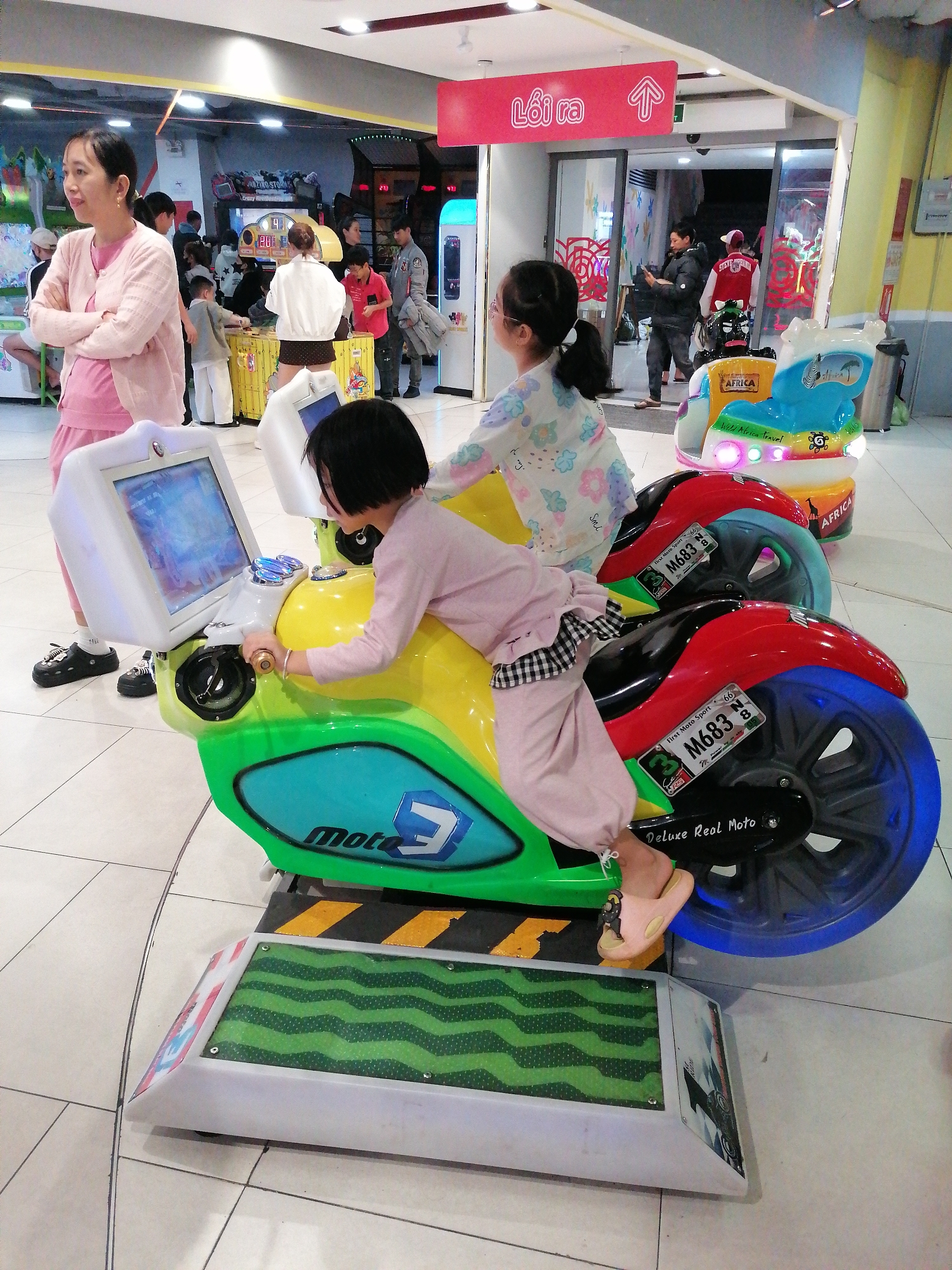 The scene is in Da Lat, Vietnam and captures an indoor play area. A young girl with short dark hair is on a yellow and green motorcycle simulator called "Moto 3" with a screen, labeled "Deluxe Real Moto." Another girl with a ponytail sits on another motorcycle simulator. An Asian woman in a pink cardigan stands to the left, observing the scene with her arms crossed. Behind the woman and girls, other people are playing games. There is signage in Vietnamese, along with other games. The floor is a neutral tile.