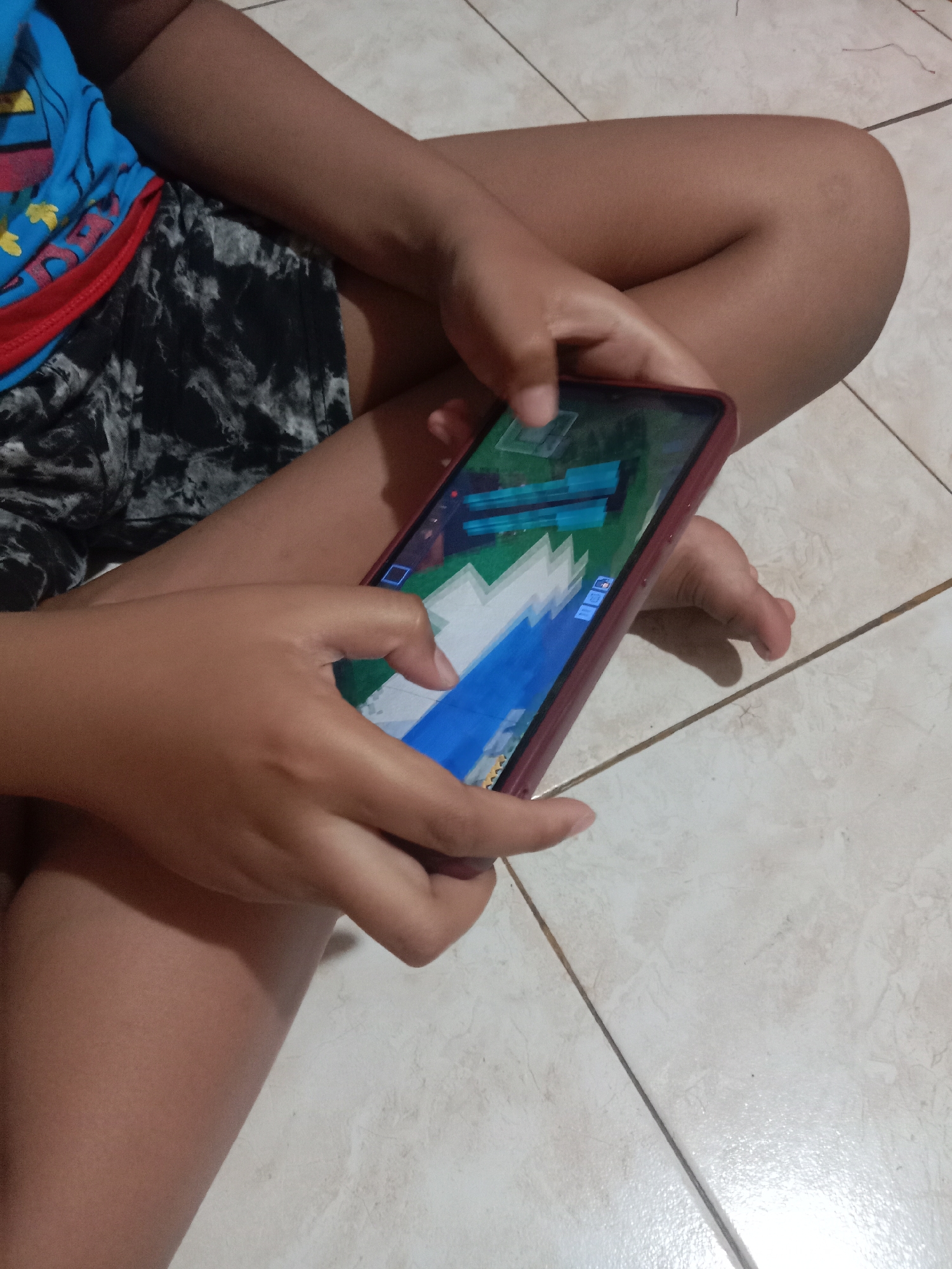 A child is sitting on a tiled floor, playing a game on a smartphone. The child is wearing a blue t-shirt with red trim and black and white patterned shorts. The child's hands are visible, holding the phone. The game on the screen appears to be a block-building game, possibly Minecraft. The background is a light-colored tiled floor with dark grout lines. The image was taken indoors, likely in a residential setting in Madiun, Indonesia.