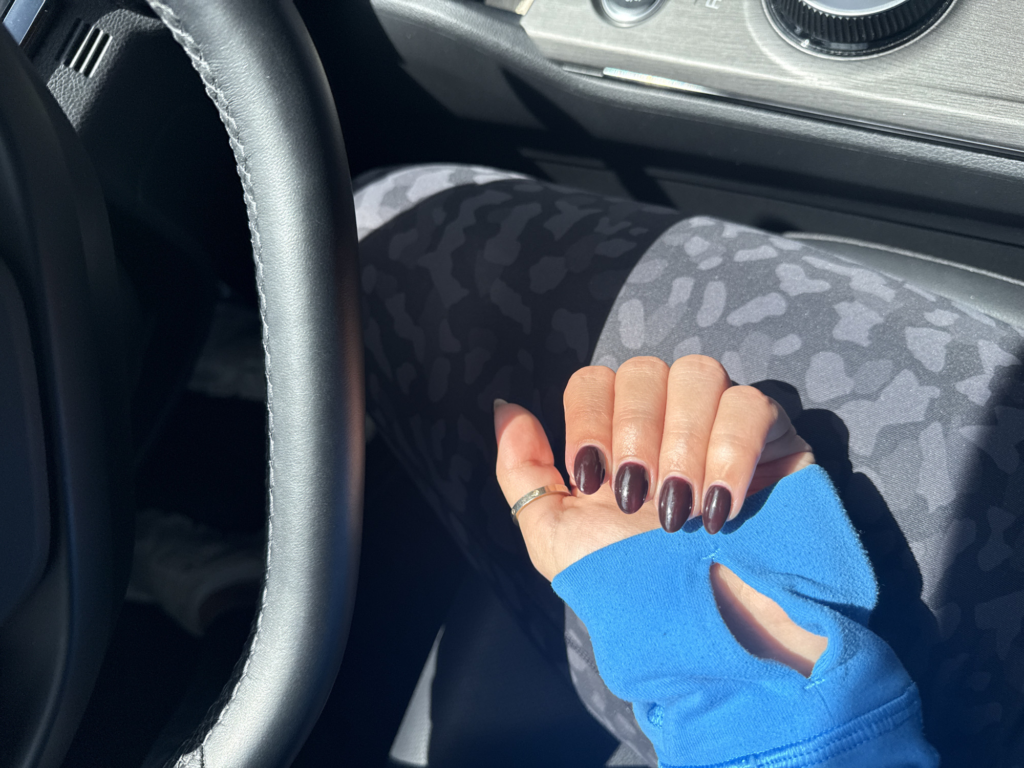 The photograph depicts a person seated in a car, indicated by the steering wheel visible on the left. The person's hand, adorned with a gold ring and dark-colored nail polish, rests on their leg, which is covered in patterned leggings. A bright blue athletic sleeve is visible around the wrist and hand, featuring a thumb hole. The car's interior elements, such as the dashboard and gear shift area, are discernible in the background. The location is not identifiable from the image.