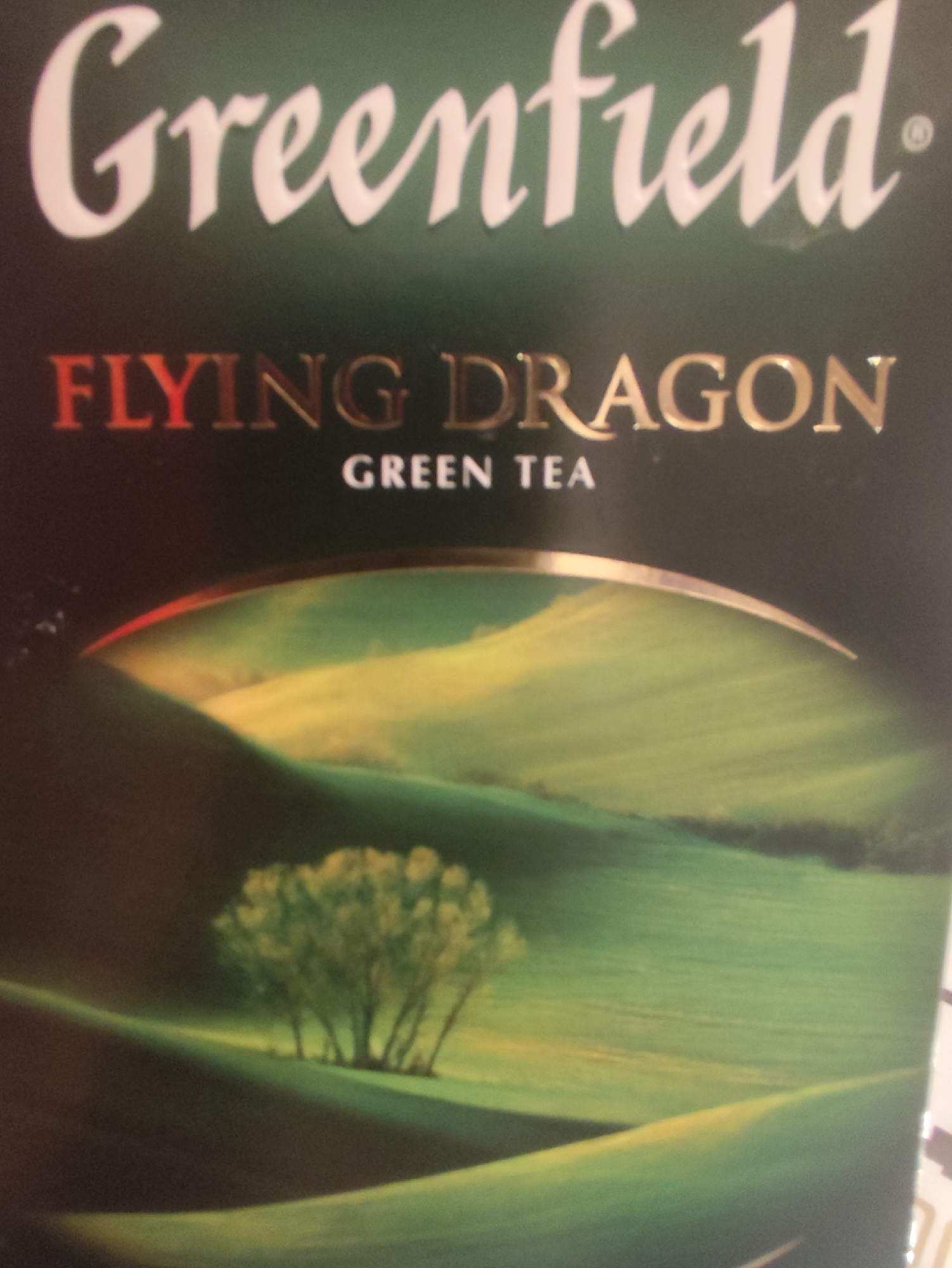 The image is a close-up shot of the packaging for "Greenfield Flying Dragon Green Tea." The packaging features a dark green background with white text for "Greenfield" in a cursive font. Below that, "FLYING DRAGON" is written in gold lettering with a red outline, and "GREEN TEA" is in white text. The lower portion of the packaging displays a picturesque landscape with rolling green hills, a yellow sky, and a cluster of trees in the mid-ground. The scene is serene and idyllic. The location in Aqbulaq Rural District, Kazakhstan, is not discernible from the image itself.