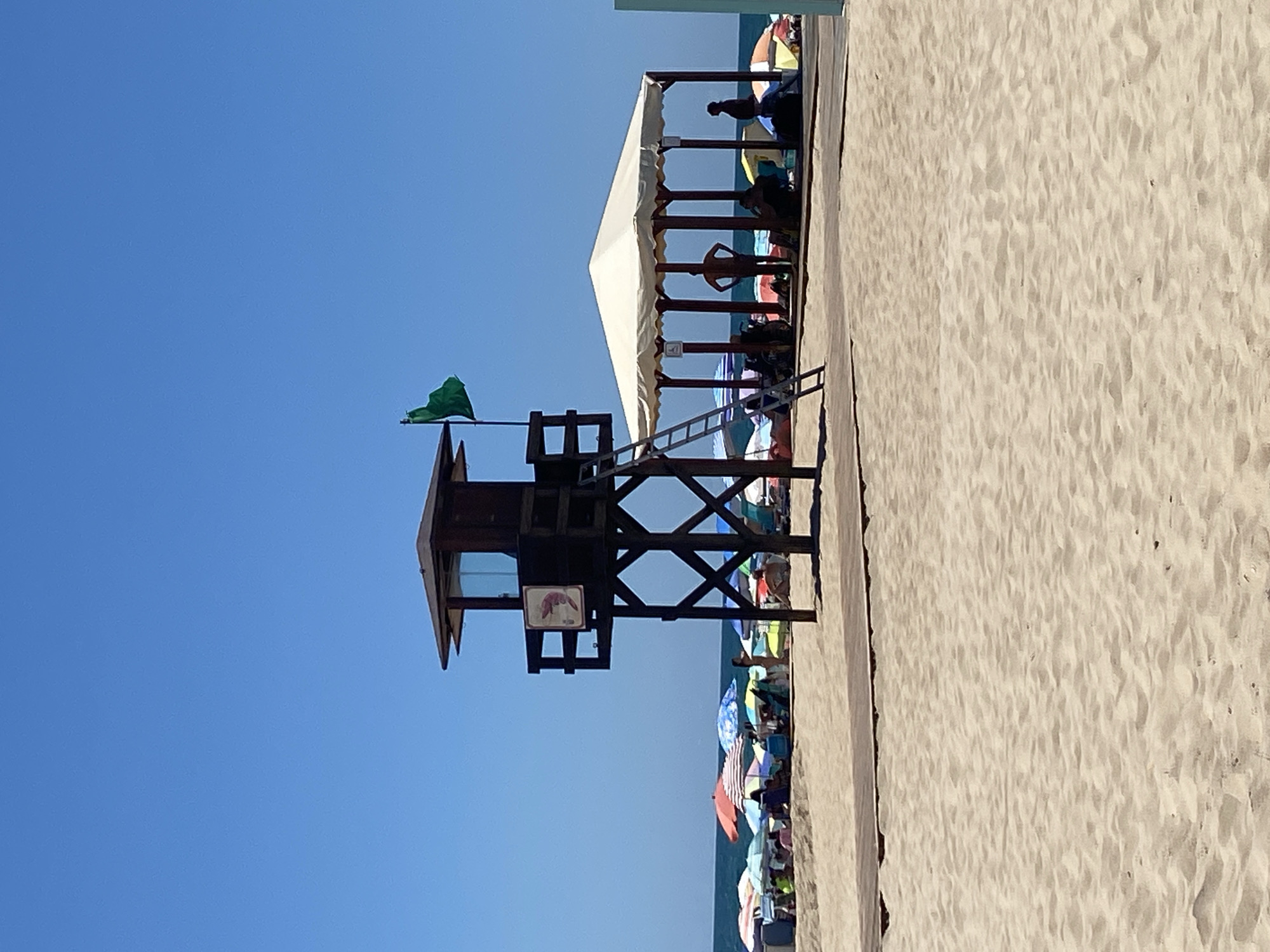 The image depicts a crowded beach scene on a bright, sunny day in Conil de la Frontera, Spain. In the foreground, a wide expanse of light brown sand is visible, with subtle ripples indicating the gentle movement of wind or water. A wooden walkway extends across the sand, leading towards the midground.
Dominating the midground is a dark brown wooden lifeguard tower, with a small enclosed cabin at the top. A ladder is attached to its side, leading up to the cabin. A green flag is flying from a pole on top of the tower. To the right of the lifeguard tower, a large, open-sided, white-covered structure with multiple posts provides shade. Several people are seen in silhouette under this structure, some appearing to be seated or standing.
Further in the background, a vibrant array of colorful beach umbrellas are spread across the sandy area, with many beachgoers seated or lounging beneath them. The sea is visible in the distance, with a clear blue sky above. The overall impression is one of a busy, recreational beach environment on a summer day.