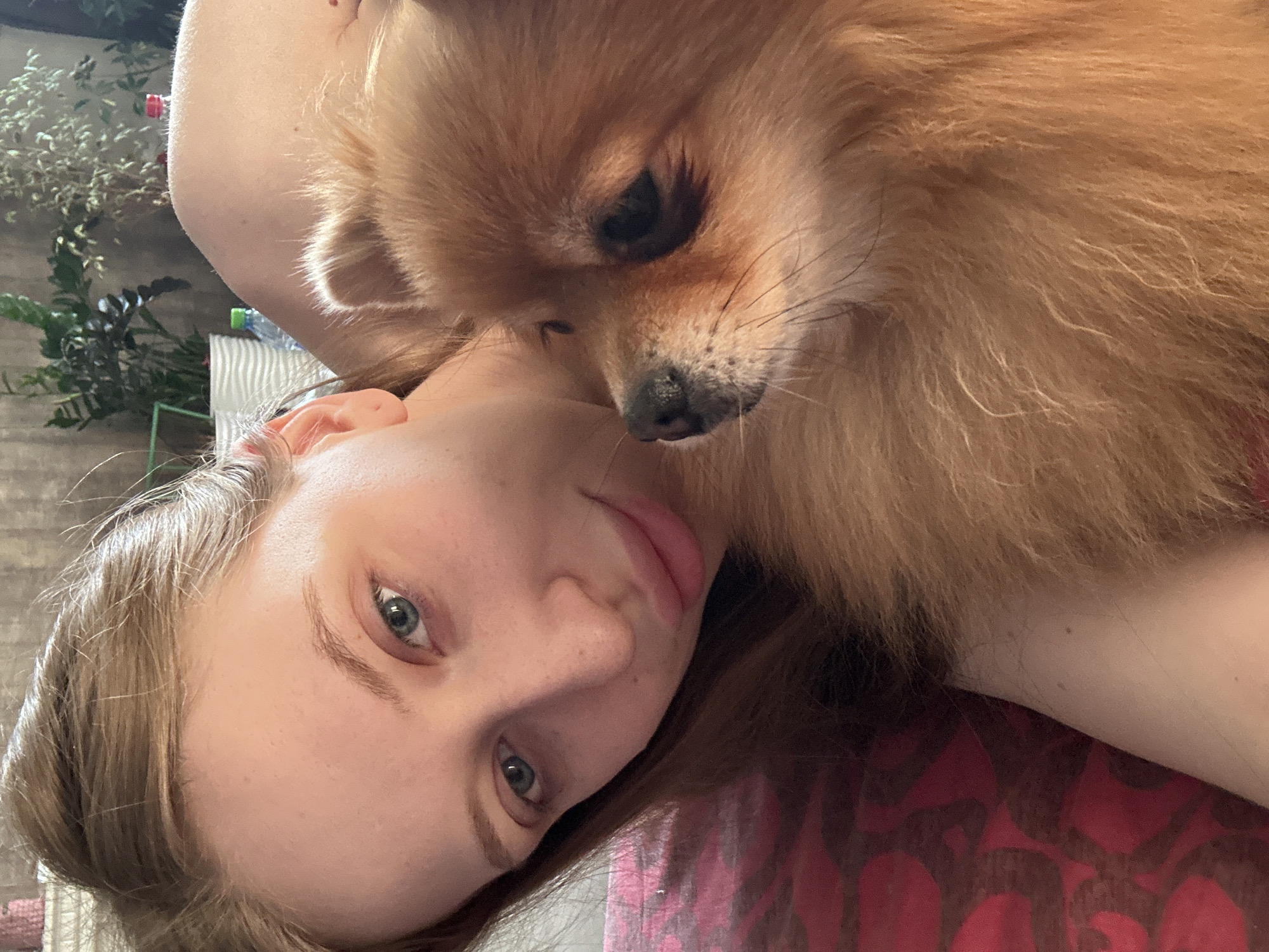 A woman is lying down with a Pomeranian dog resting its head near her face. The woman has fair skin, light brown hair, and blue eyes, and is looking at the camera. The dog is orange-brown. The background includes a floral-patterned surface. The image was taken in Chaikovsky, Russia.