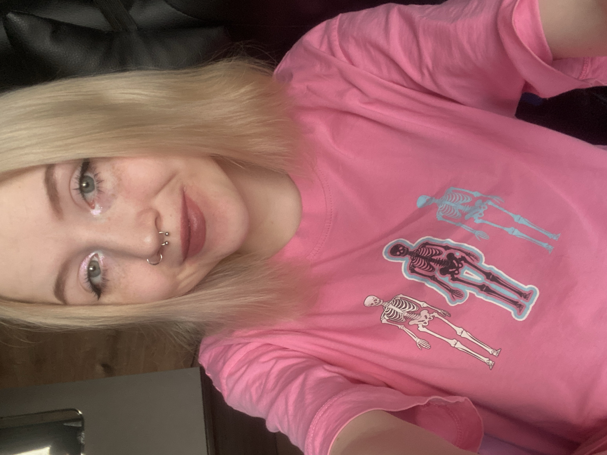 A young female subject, with fair skin, shoulder-length blonde hair, and blue-green eyes, is depicted in a close-up, front-facing orientation. She wears light-colored eyeshadow, black eyeliner, and reddish-brown lipstick. Her nose features two piercings: a hoop in the right nostril and a horseshoe barbell in the septum. The subject is wearing a bright pink, short-sleeved, crew-neck t-shirt adorned with three skeleton graphics on the front; from left to right, these include a white skeleton, a dark skeleton outlined in light blue, and a partially visible light blue skeleton. The left arm of the subject is visible, bent at the elbow, indicative of a self-portrait (selfie) capture. The background is largely out of focus, revealing indistinct elements: a blurry light-colored vertical surface and a reflection on the far left, a warm-toned wooden surface in the center-left, and a dark, textured surface, possibly a leather chair or car seat, on the right. The specific location within Izhevsk, Russia, is not discernible from the provided visual information.