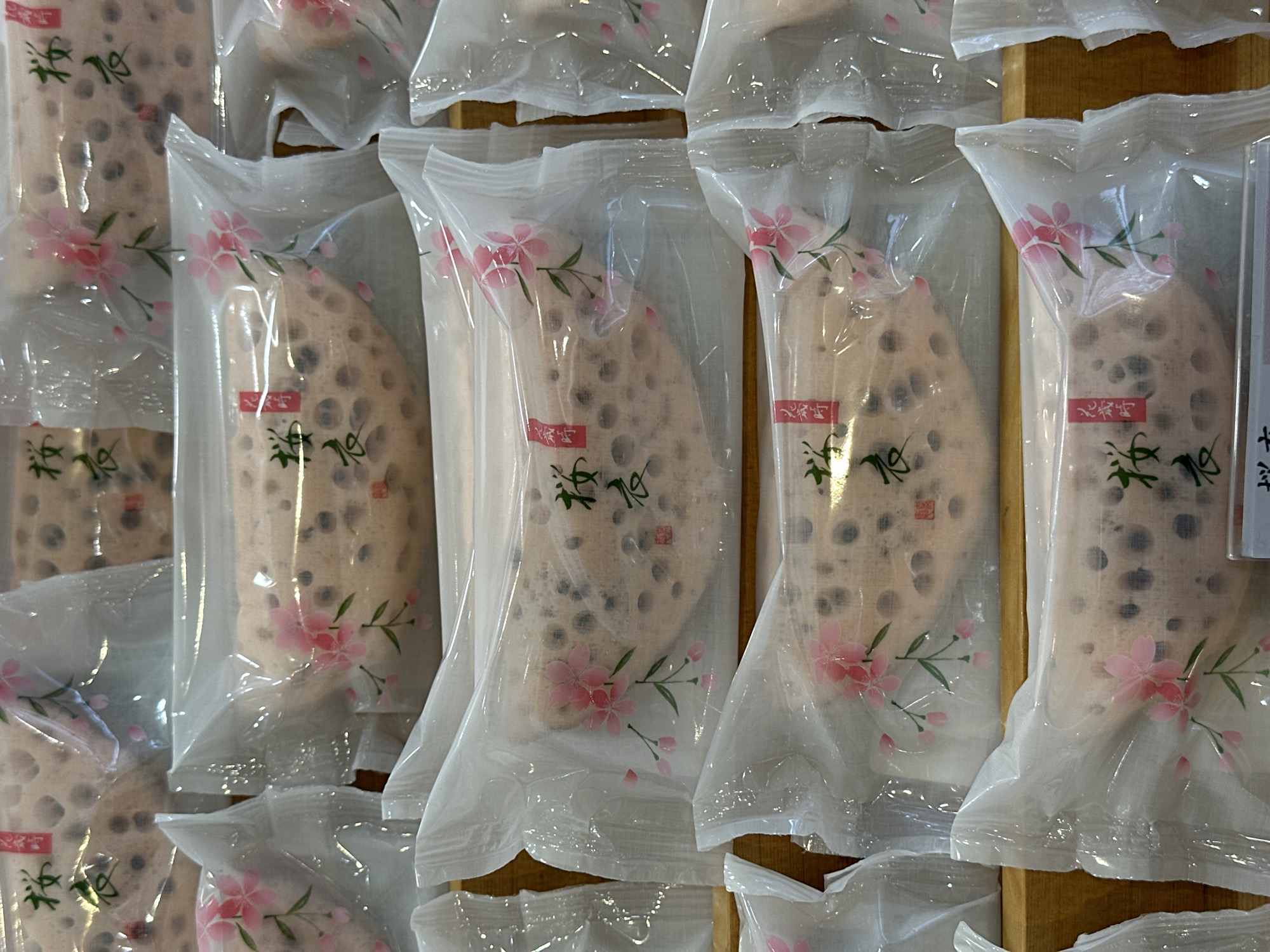 The image shows multiple packages of what appears to be lotus root snacks, each individually wrapped in clear plastic. The packaging is decorated with a pink floral design. The snacks are arranged in a row, likely on a shelf or display. The location is not discernible from the image, and there is no information to suggest that the location is in London, United Kingdom.