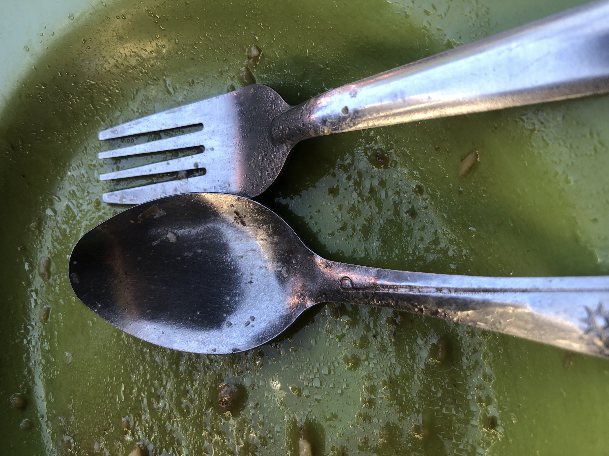 A fork and spoon are resting on a green plate with remnants of a meal. The utensils and plate are wet and appear to be lightly used. The image is taken from a top-down perspective. This scene is likely from a restaurant or home kitchen in Surabaya, Indonesia.
