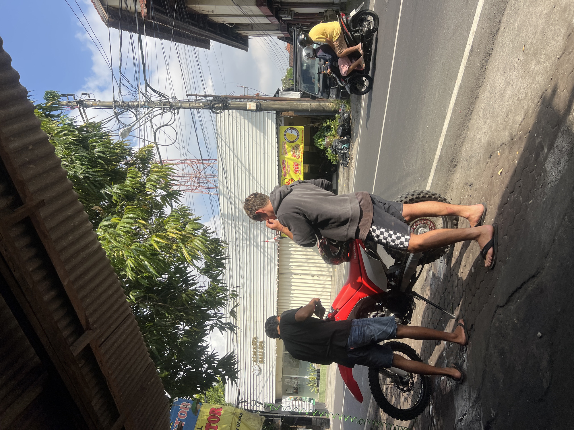 The image captures a street scene in Denpasar, Indonesia, possibly near a shop or service area, as indicated by the corrugated metal overhang providing shade. Two men are present. One man, wearing a black t-shirt and denim shorts, stands beside a red dirt bike, appearing to be attending to it. The other man, dressed in a grey hooded sweatshirt and checkered shorts, stands slightly behind and to the right, holding a phone to his ear. A red dirt bike is parked on the textured pavement.
In the background, a white building with vertical slats is visible, bearing a sign with Indonesian text and a logo that appears to be "ALMISTA BALI SPA." Adjacent to this is a smaller establishment with a yellow sign featuring text and images, one of which appears to be a cartoonish curry dish, with the text "SCC" prominently displayed. Further down the street, a black van is parked, and a motorcycle carrying two people, a driver and a passenger, is in motion, traveling on the asphalt road. The road is marked with white lines. Overhead, numerous power lines and a utility pole are visible against a bright blue sky with scattered clouds. The environment suggests a warm, sunny day.