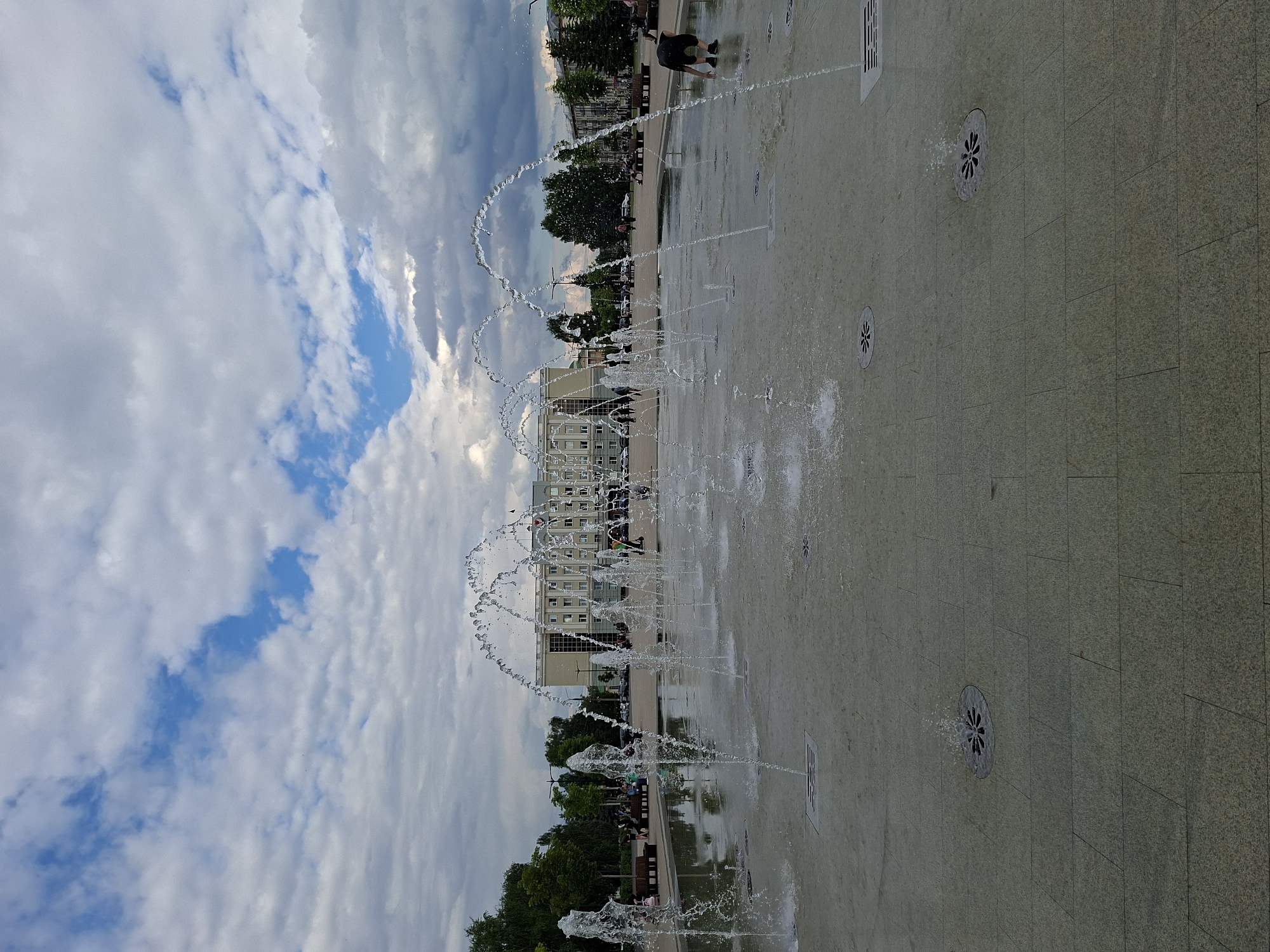 The image depicts a public square in Izhevsk, Russia, featuring a modern fountain system with multiple jets of water spraying in arcs. The square is paved with stone tiles and has several drainage grates. In the background, a large, classical-style building with a prominent central section and colonnades stands behind a line of trees. Several people are visible in the background, some seated on benches and others walking. One person, a child or young adult, is crouched near the fountain in the foreground, appearing to interact with the water. The sky is partially cloudy with patches of blue.