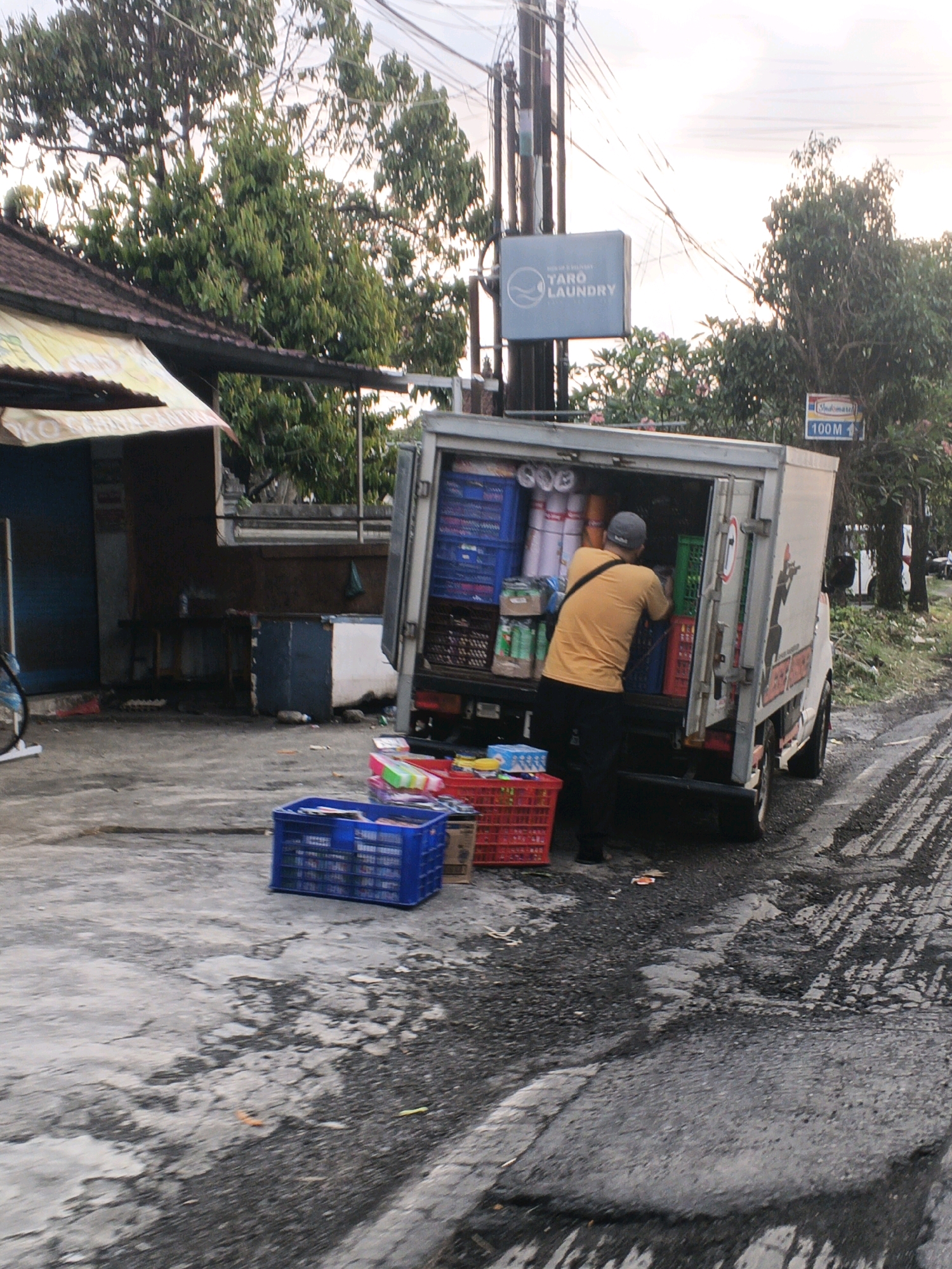 In Sukawati, Indonesia, a man wearing a yellow t-shirt and black pants is seen unloading goods from the back of a white delivery truck. The truck is parked on a wet, cracked asphalt road with tire tracks visible. The rear of the truck is open, revealing an assortment of items, including blue and green plastic crates, white rolls of material, and other packaged goods. In the foreground, several red and blue plastic crates are on the ground, some filled with items. Behind the truck, a sign for "TARO LAUNDRY" is visible on a pole, along with a smaller sign indicating "Indomaret 100 M". The scene is set against a backdrop of lush green trees and a building with a dark roof. The lighting suggests it is either early morning or late afternoon.