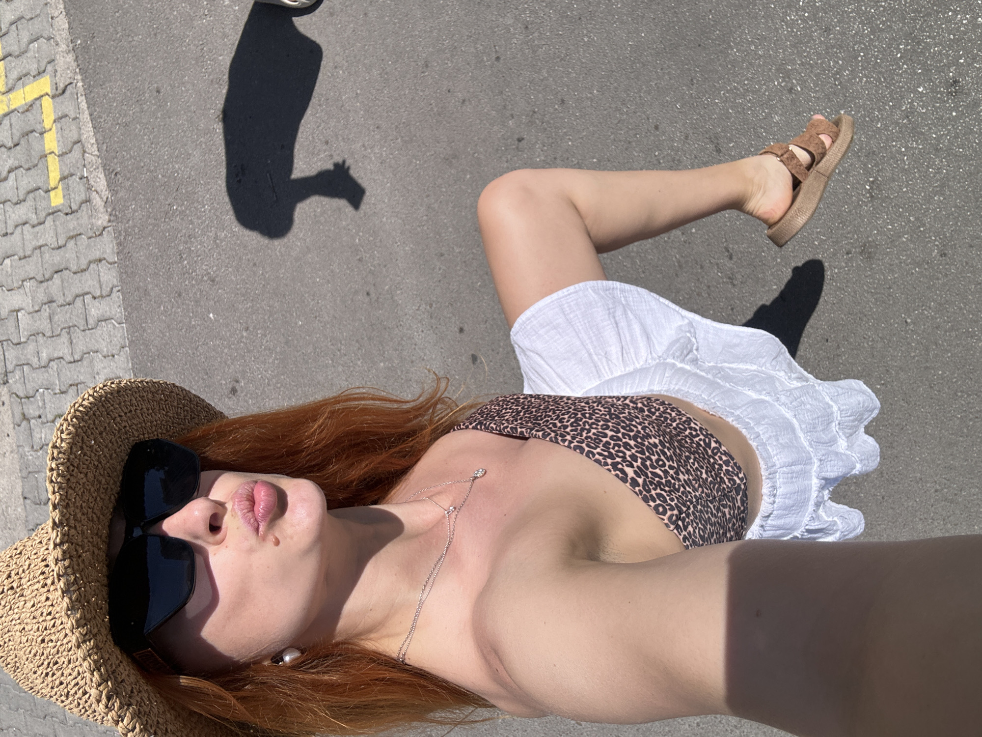 A person is lying on a paved surface, possibly a street, in what appears to be Budva, Montenegro, judging by the yellow pavement markings which are common in the city. The person is wearing a straw hat, dark sunglasses, and a leopard-print top, along with a white skirt. They are wearing sandals on one foot. The person is facing upward towards the camera, and their arm is extended towards the camera. A shadow of someone or something else is visible nearby on the pavement.