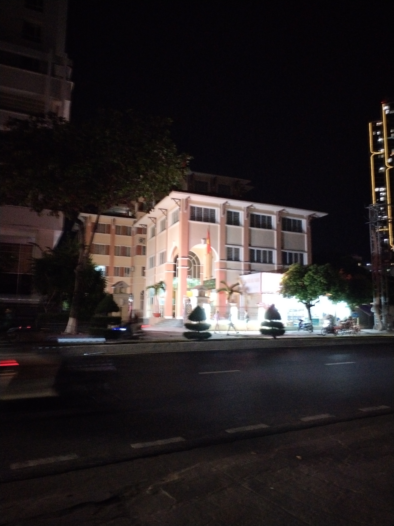 The image depicts a nighttime scene in Bac Nha Trang Ward, Vietnam, featuring a building with illuminated windows and architectural details, possibly a government or educational institution, given the presence of a flag. The building is situated alongside a street with visible road markings and some traffic, indicated by motion blur from vehicles. Several trees and manicured bushes are present in front of the building, with some individuals walking on the sidewalk and a few motorcycles parked or in motion. The overall atmosphere is urban and illuminated by artificial light against a dark night sky.