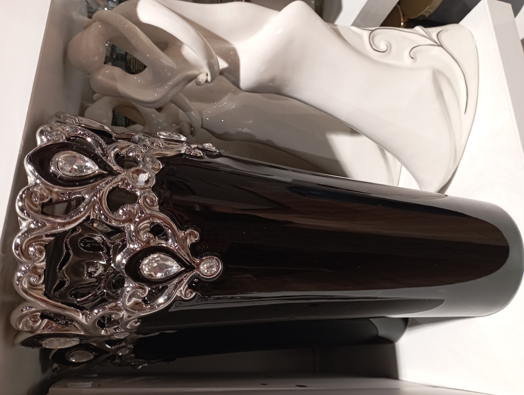 The image shows a black vase with ornate silver and rhinestone detailing at its rim. The vase is tall and cylindrical, with a glossy finish. It is positioned in the foreground, slightly to the left of center. Behind the vase, to the right, are two white ceramic sculptures that appear to be abstract figures in an embrace. They are smooth and flowing in design, with subtle decorative swirls on the base of the right sculpture. The objects are placed on white shelves, suggesting they are part of a retail display. The background is indistinct, with hints of other objects and shelving visible. The lighting is bright, creating reflections on the glossy surfaces of the vase and sculptures. The scene is indoors, likely in a store or showroom. There are no people visible in the image. This image was taken in Katowice, Poland, at Galeria Katowicka.