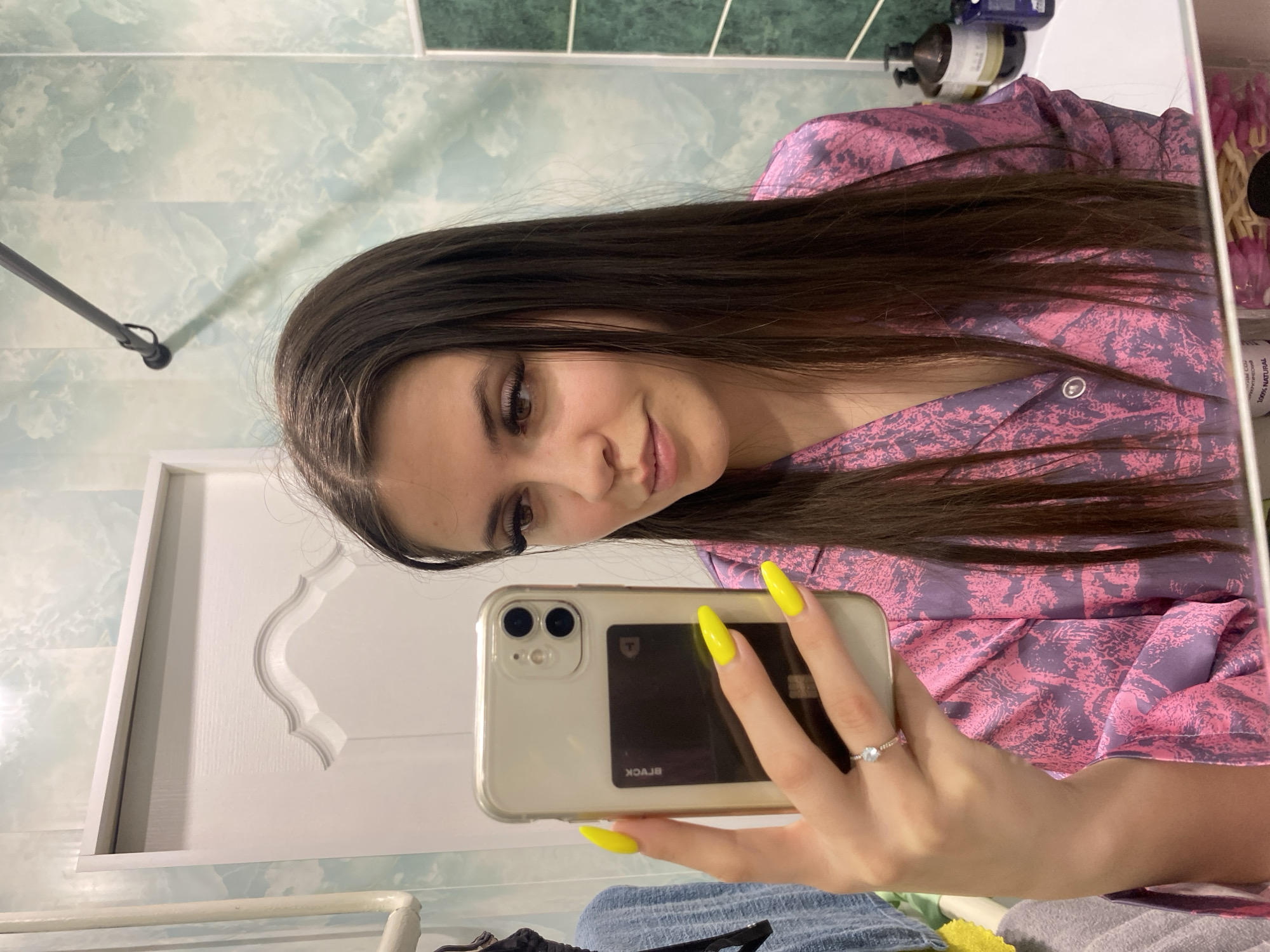 A young woman with long, dark hair is taking a selfie in a bathroom. She is wearing a pink and purple patterned top. Her nails are painted bright yellow. She is holding a white iPhone in her right hand. The bathroom has white and green patterned tiles on the walls and a white door with decorative molding. The location is likely in the city of Chaikovsky, Russia.