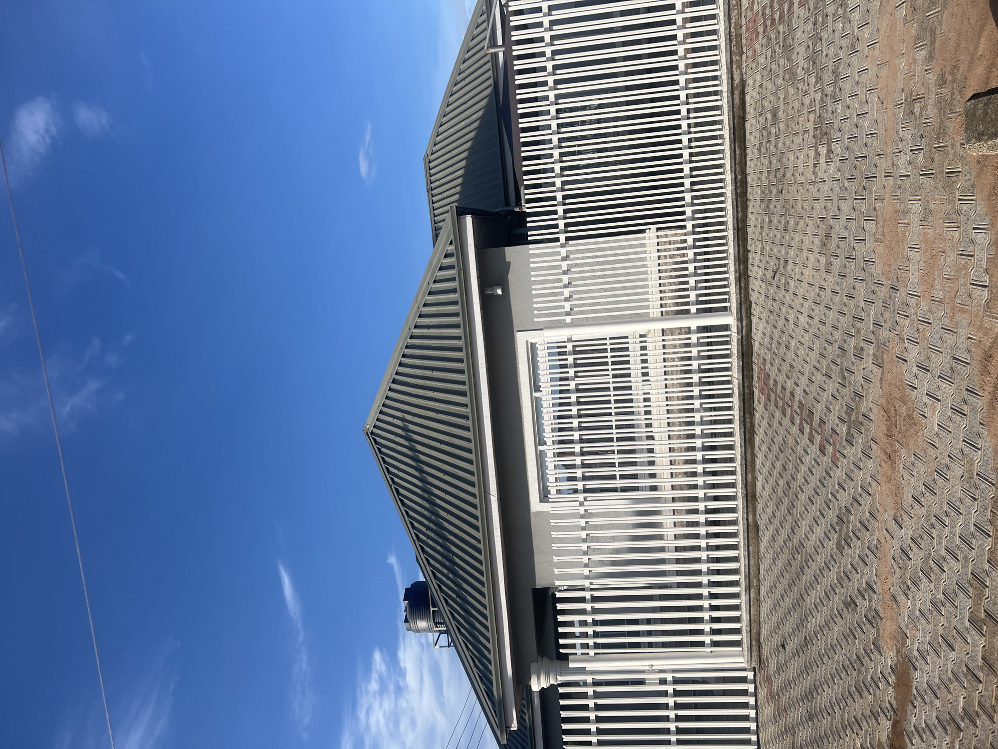 A vertically oriented photograph captures a modern building under a clear blue sky, likely in Jalingo, Nigeria. The scene is illuminated by bright sunlight, casting strong shadows and indicating a midday or early afternoon setting.
The building, positioned to the left and center, features a multi-faceted dark gray corrugated metal roof with visible gutters. A black cylindrical water storage tank is situated on one of the roof sections. The upper part of the building's facade is characterized by an extensive white slatted structure, appearing as a protective screen or an architectural element for a balcony or window area. Below this, the building wall is a light gray.
To the right of the building, a paved ground area is visible. It is made of interlocking bricks or tiles, displaying a geometric pattern with shades of gray and reddish-brown. This surface extends from the foreground towards the background.
The sky occupies a significant portion of the image, showcasing a vibrant, clear blue with only a few sparse, wispy white clouds. A thin, dark power line traverses the upper left section of the frame against the blue sky. There are no people visible, and no specific activity or event is taking place. No discernible text is present in the image.