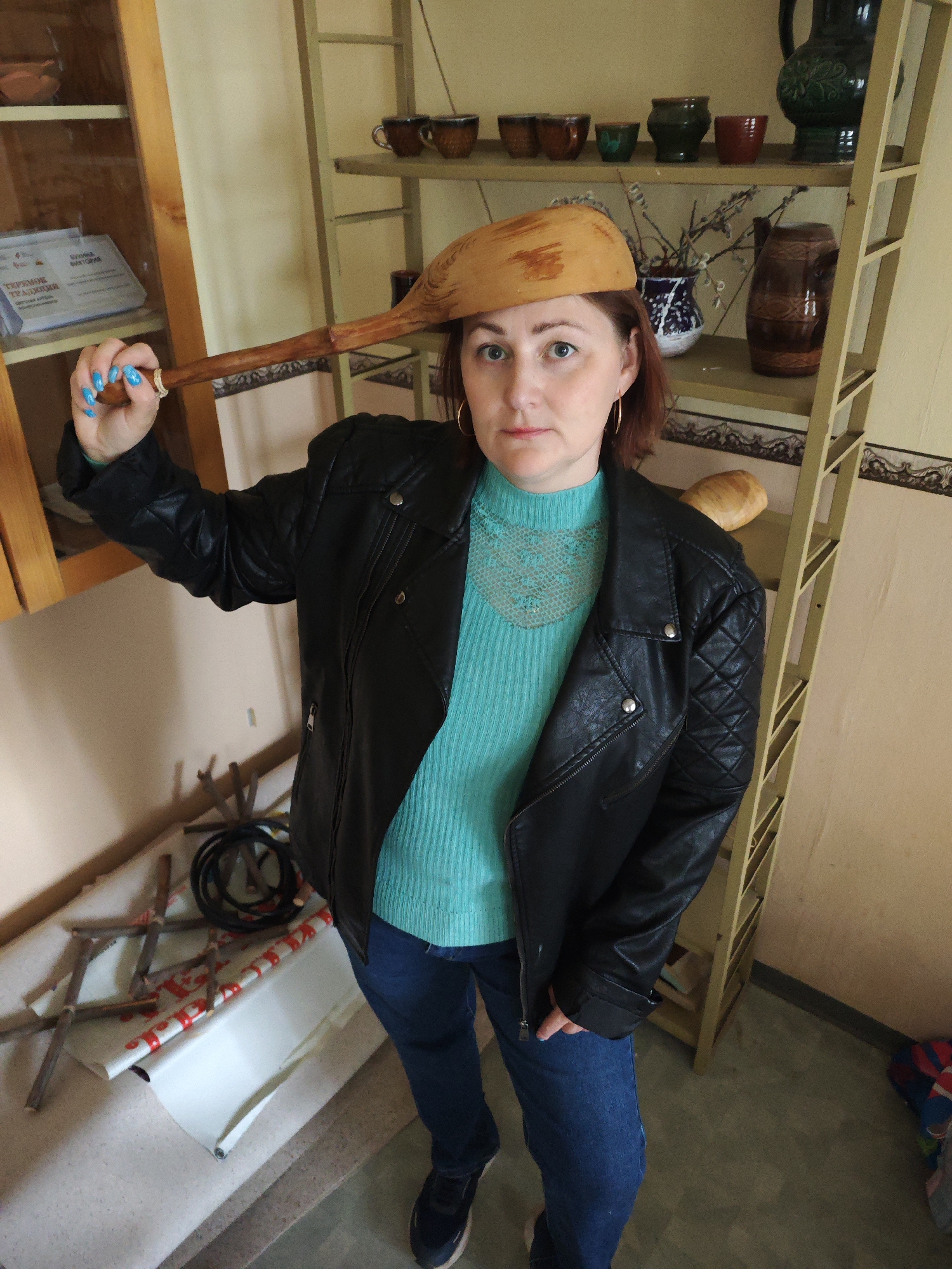 This full-shot portrait captures a woman posing indoors, likely within a cultural center, museum, or shop in Markovskii, Russia, dedicated to traditional crafts. She is depicted in her 30s or 40s, with light brown hair and direct, light-colored eyes. Her attire consists of a black faux leather motorcycle-style jacket, a ribbed teal turtleneck sweater featuring a subtle lace detail at the neck, blue jeans, and dark athletic shoes. Notable accessories include golden hoop earrings and vibrant blue nail polish.
A distinctive element is the large, light-colored wooden spoon or similar utensil she playfully balances on her head, holding its handle with her left hand. Her expression is calm and direct as she gazes towards the viewer.
The background offers context: to her left, a light wooden display cabinet with glass doors houses several ceramic bowls and a framed document. Partially visible Cyrillic text on this document clearly shows "ТРАДИЦИЯ" (Tradition), confirming the cultural theme. To her right, a taller, narrow shelf unit, possibly made of light wood or metal, is filled with an assortment of rustic pottery – including dark brown cups, green and red pots, and a large green jug – alongside a vase of bare branches and a loaf of bread. The walls are a plain, light hue. On the floor in the foreground, various items are visible: haphazardly arranged wooden sticks, a coiled black cable, and a rolled-up white banner or poster with red designs or text. The overall scene evokes an environment rich in local heritage and artisanal products.