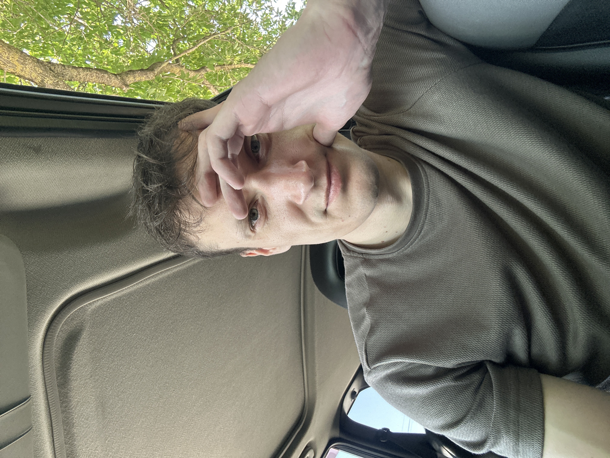 A man is sitting inside a car. The car's sunroof is open, revealing green foliage from a tree overhead and a bright sky. The man is looking towards the camera, with his left hand resting on his forehead and his right hand on his cheek. He is wearing a taupe, textured long-sleeved shirt. The interior of the car is visible, including the headliner and a portion of a car seat. The scene appears to be located outdoors, near a tree, possibly in the village of Makiv, Ukraine, given the context.