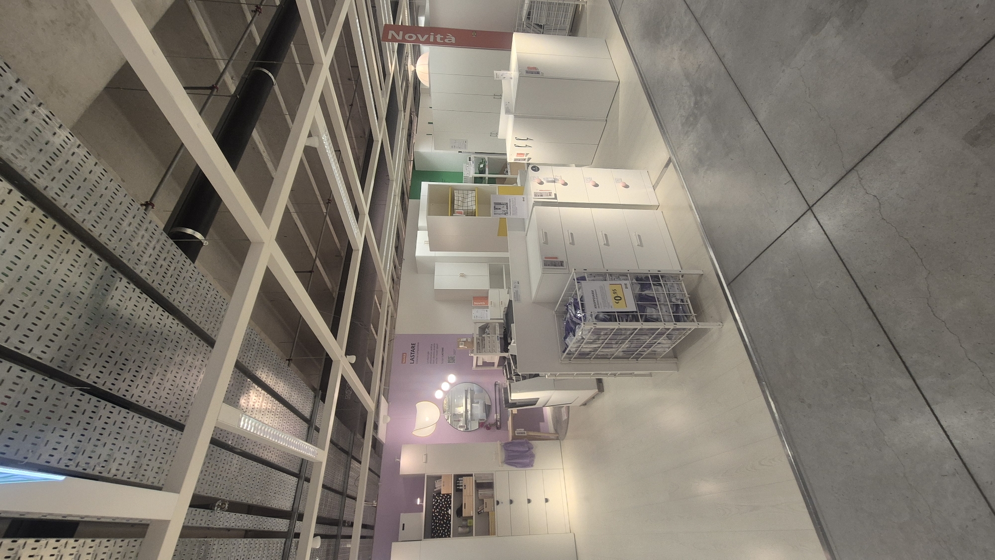The image depicts the interior of a retail store, specifically an IKEA store, as indicated by the "Novità" sign and the characteristic product displays. The scene showcases various furniture items, primarily white cabinets and drawers, arranged in a showroom-like setting. In the foreground, a wire basket filled with packaged goods is visible, with a price tag of €0.95. Behind this, a set of white drawers and cabinets are displayed. The background features more furniture, including larger wardrobes and shelving units, some with yellow accents. A purple wall serves as a backdrop to a vanity area with a mirror and desk. The ceiling is industrial with exposed conduits and lighting fixtures. The floor transitions from a light wood-like laminate to concrete. The store appears to be located in Baronissi, Italy, given the presence of the "Novità" sign, which translates to "New Arrivals" in Italian. There are no people visible in the image.