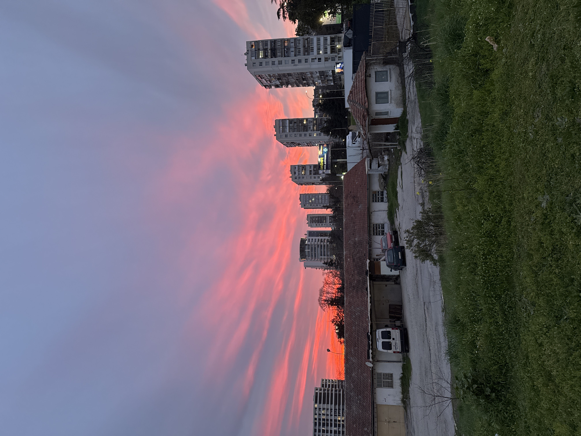 The image shows an urban scene in Varna, Bulgaria, at dusk or dawn. The sky is filled with pink and purple-streaked clouds. Several tall apartment buildings are visible in the background, indicating a dense urban environment. In the foreground, there are lower-level buildings, possibly houses or shops, with tiled roofs. A narrow road or alley runs alongside these buildings, with a white van parked on it. Vegetation, likely grass and bushes, grows along the side of the road. The overall impression is of a residential area on the outskirts of a larger city, captured during a colorful sunrise or sunset.