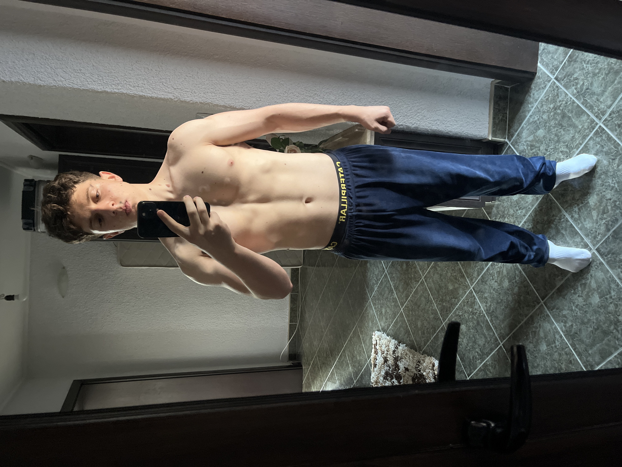 An indoor mirror selfie depicts a young male, appearing to be in his late teens or early twenties, standing shirtless in a room located in Zlatograd, Bulgaria. He has short, curly dark hair and light stubble on his upper lip and chin. Visible on his skin are multiple moles and distinct patches of lighter hypopigmentation on his left shoulder and upper chest. He holds a black smartphone in his right hand, obscuring the lower half of his face, as he captures his reflection. The individual wears dark blue velvet or velour sweatpants with a yellow "CATERPILLAR" brand waistband, which is reversed due to the mirror reflection, and white crew-length socks.
The room features dark green-grey tiled flooring with white grout lines forming a grid pattern, and off-white textured walls. A dark brown door, slightly ajar with a black handle, is visible on the left, with another similar door frame visible on the right. A small, shaggy rug with brown and white patterns lies on the floor to the left. A thin white electrical cable runs along the far wall near the floor. Reflected in the mirror behind the individual is a bare light bulb fixture hanging from the ceiling and a light-colored object, possibly a cushion or headboard, against the wall. To the subject's right, a decorative element, possibly a vase containing a rose, is reflected on a narrow surface.