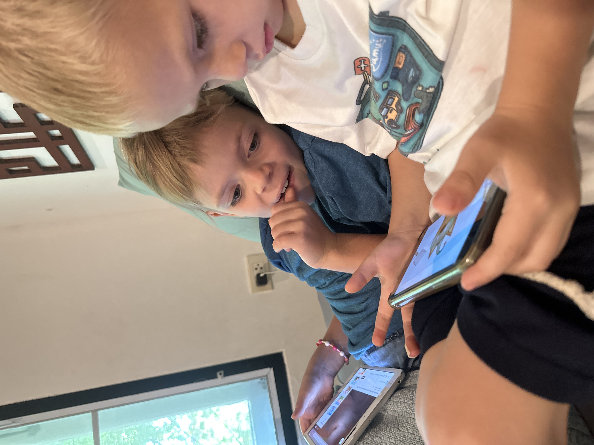 Two young boys are indoors, likely in a residence. One is wearing a white t-shirt with a cartoon design, while the other wears a blue shirt. They are both intently focused on a smartphone and a tablet, suggesting they are watching videos or playing a game. The scene includes a window, a wall outlet, and part of a patterned wall decoration. The presence of electronic devices, along with the children's attire, indicates a relatively modern setting. The location is ตำบลเกาะพะงัน, Thailand.