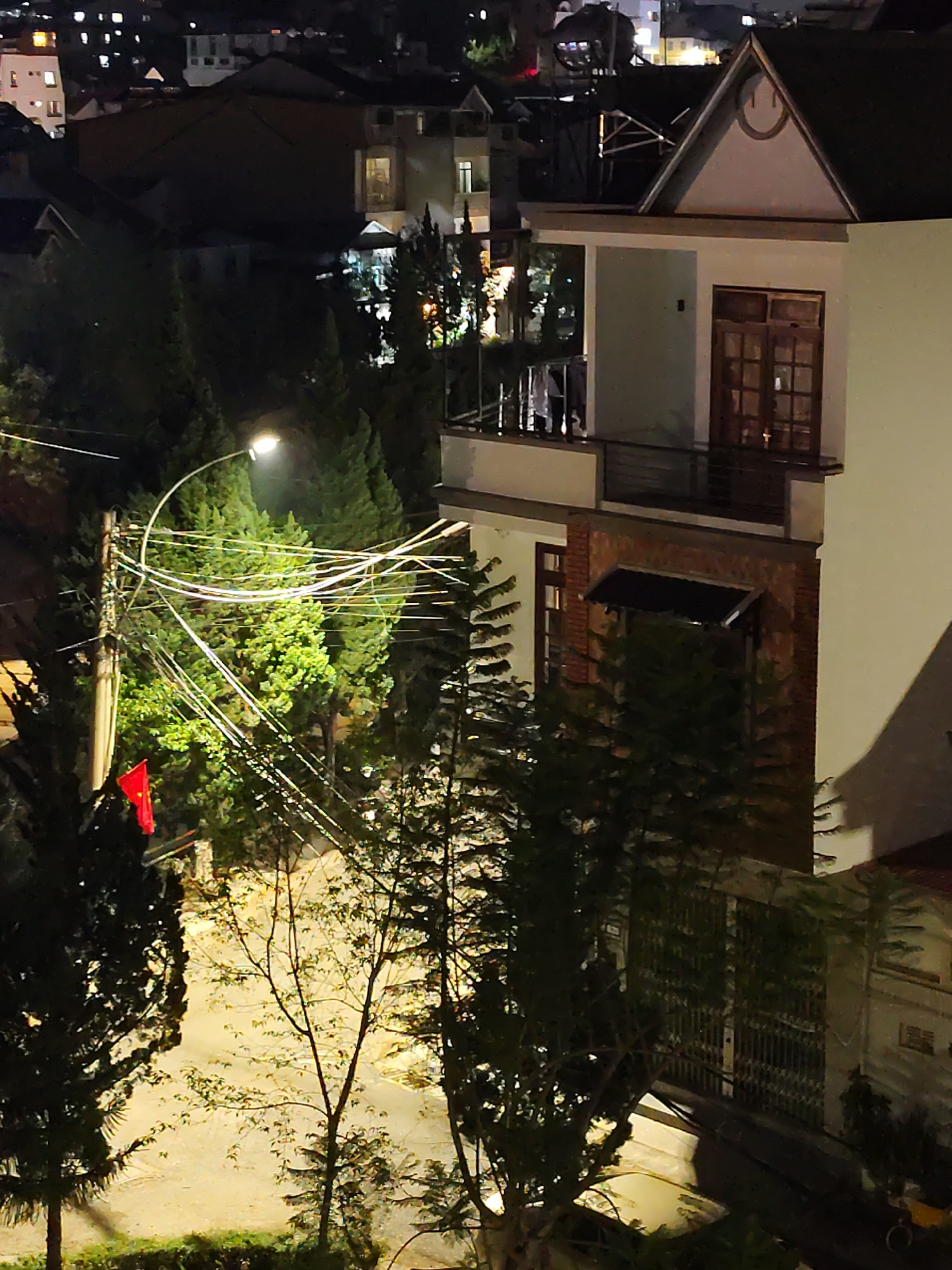 The image is a nighttime view in Da Lat, Vietnam, showing a two-story building with a sharply angled gable roof. The upper portion of the building is white, while the lower portion is brick. A balcony with a metal railing is visible on the second story. Dense evergreen trees and other vegetation partially obscure the building and the street below. Streetlights cast a bright, yellowish light on the road and surrounding foliage. A power pole with a red flag attached is visible to the left, and numerous electrical wires crisscross in front of the trees. Other buildings are visible in the background, with lights indicating nighttime activity.