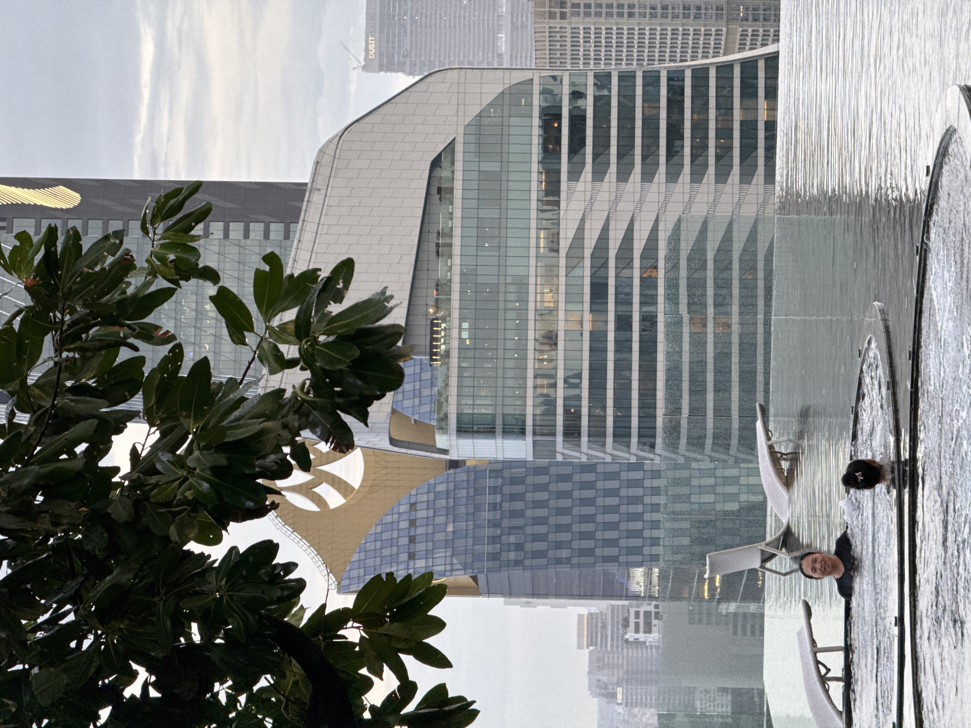 The image captures a cloudy day in Bangkok, Thailand, from a rooftop perspective. In the foreground, an infinity pool overlooks a cityscape dominated by modern skyscrapers. Two individuals are visible in the pool. A woman with dark hair, wearing a black shirt, is positioned in the center, looking towards the camera with a smile. Behind her, another person with dark hair pulled back is visible from the shoulders up. In the background, a cluster of contemporary buildings with distinct architectural styles pierce the sky. One building features a striking gold-colored curved structure at its apex. Trees with dark green leaves frame the upper left portion of the image. The overall scene suggests a luxurious urban oasis amidst the bustling city.