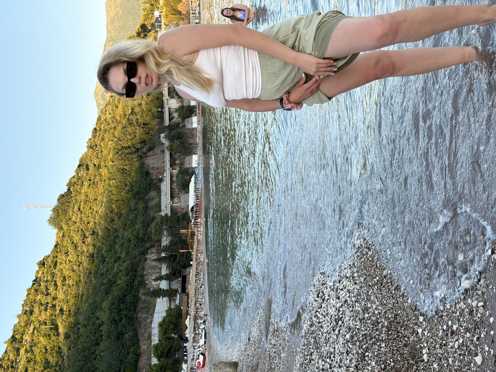 In Budva, Montenegro, a blonde woman with long wavy hair, wearing large black sunglasses, a white tank top, and olive green shorts, stands knee-deep in the calm sea water near a pebbled beach. She is posing for a photo, lifting her shorts slightly with her hands. In the background, another woman in a blue bikini is visible in the water. The beach is lined with rows of lounge chairs and umbrellas, and a forested hillside rises behind the beach, with a road and some structures visible on its slope. The sky is clear and blue, and the sun is casting a warm glow on the scene.