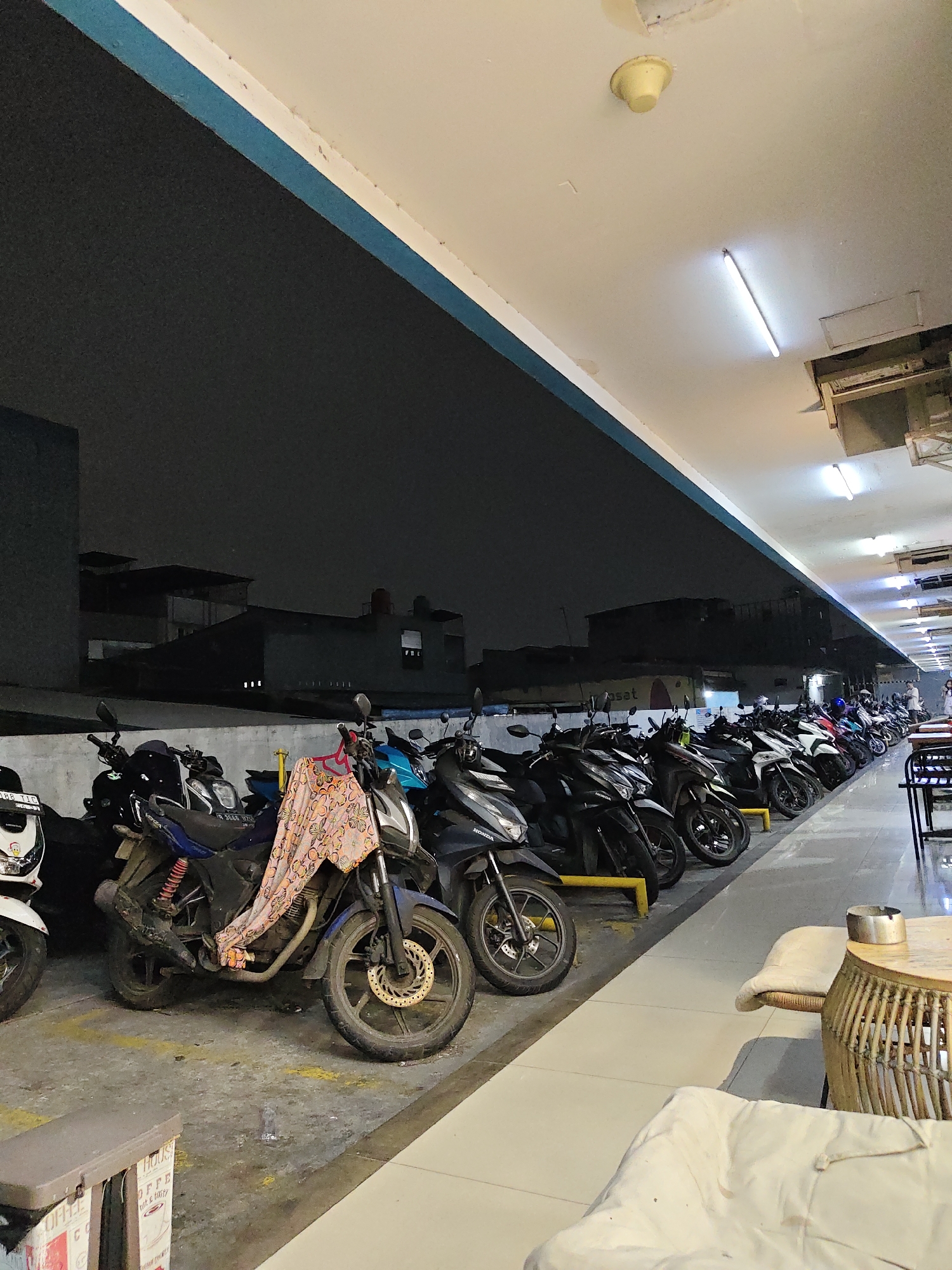 A nighttime outdoor parking lot in North Jakarta, Indonesia, is filled with numerous motorcycles. The parking spaces are marked with yellow lines on a concrete floor. A few rows of motorcycles are parked, facing left, with some of them covered by patterned cloth. In the foreground, a woven round table with an ashtray and two cushioned chairs are visible, suggesting a seating area. Above, the ceiling is white with fluorescent lighting, and a circular yellow smoke detector is mounted. The background shows a dark sky and silhouettes of buildings. The scene is lit by artificial lights from the ceiling and possibly streetlights.