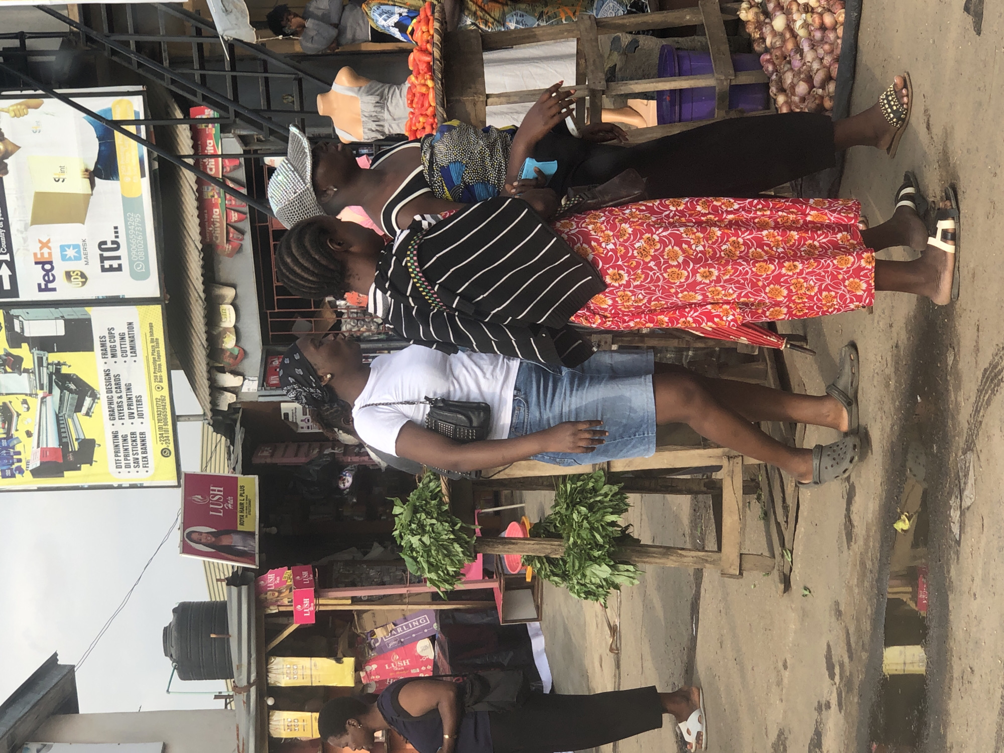 The image depicts a street scene in Lagos, Nigeria, likely an open-air market or commercial area. In the foreground, three women are walking. The woman on the left wears a dark blue top and black pants, with white sandals. The woman in the middle is wearing a white t-shirt, denim shorts, and grey Crocs. She is carrying a black purse. The woman on the right wears a black and white striped top with a colorful wrap around her waist, a red floral skirt, and patterned flip-flops. She is carrying a small blue object.
Behind the women, there are market stalls. One stall has a display of leafy green vegetables on a wooden stand. Another stall features a pile of red tomatoes on a wooden surface, and further to the right, a large quantity of onions are visible. Several signs are displayed, including one for "LUSH HAIR," advertising hair products, and another large banner with graphics and text related to printing services and shipping companies like FedEx and UPS. The environment appears to be dusty and the ground is uneven, with puddles of water visible in the foreground. The overall impression is a vibrant, bustling urban environment in Lagos.