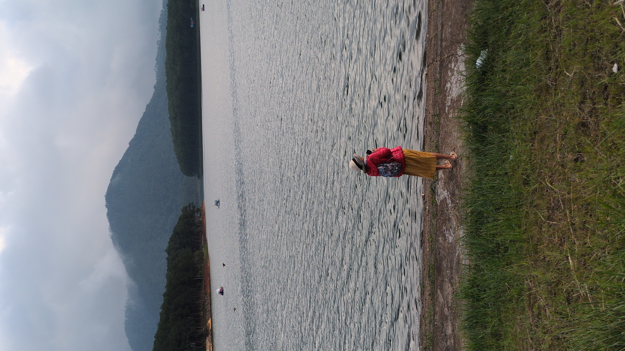 A woman stands at the edge of a large body of water, facing away from the camera. She is wearing a red jacket, a yellow skirt, and a light-colored hat. A backpack with a colorful pattern is on her back. She appears to be holding a phone or camera in her right hand. The water is calm with ripples, and a line of trees forms a dense forest along the far bank, which transitions into a misty mountain range in the background. Several small boats are visible on the water, including paddle boats. The foreground consists of a concrete path next to a patch of grass, with some scattered white debris. This scene is likely Tuyen Lam Lake in Da Lat, Vietnam, known for its scenic beauty and recreational activities.