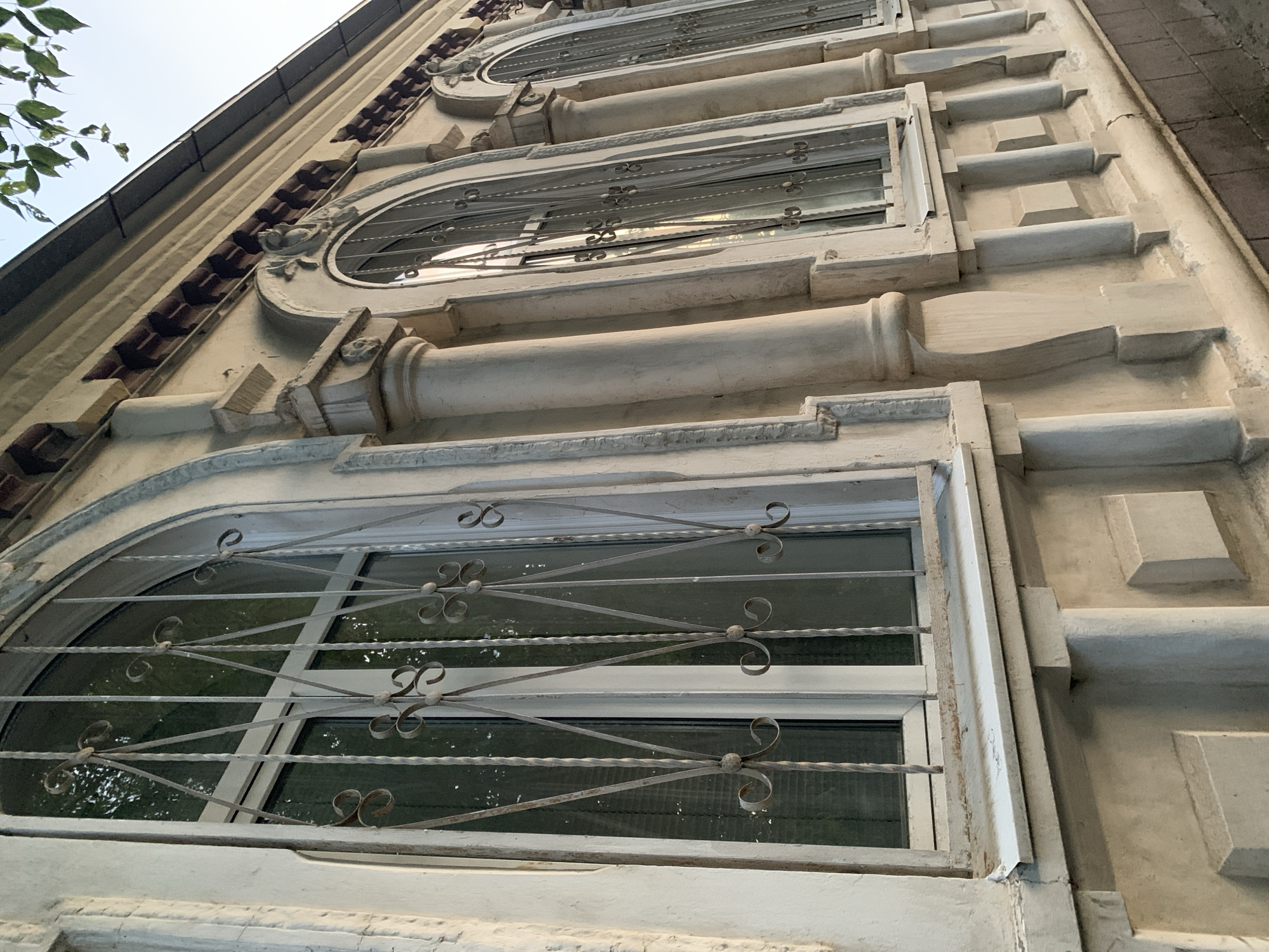 The image shows a close-up, low-angle exterior view of a building facade in a city. The facade is constructed of light-colored material, likely stucco or stone, with decorative architectural elements. Several arched windows are visible, each fitted with ornate metal security bars. The windows appear to have blinds or coverings behind the bars.
The architectural details include pilasters and cornices, suggesting a classical or neoclassical style. The building's color is a pale, creamy hue, and some areas show signs of age and wear, such as minor discoloration or chipping.
The lighting suggests it is either early morning or late afternoon, with long shadows cast by the architectural features. Above and to the right of the building, a few green leaves from a tree are visible against a pale sky. The ground level is not clearly depicted, but a hint of brickwork is visible at the very bottom right, suggesting a walkway or paved area. There are no people or vehicles in the frame.