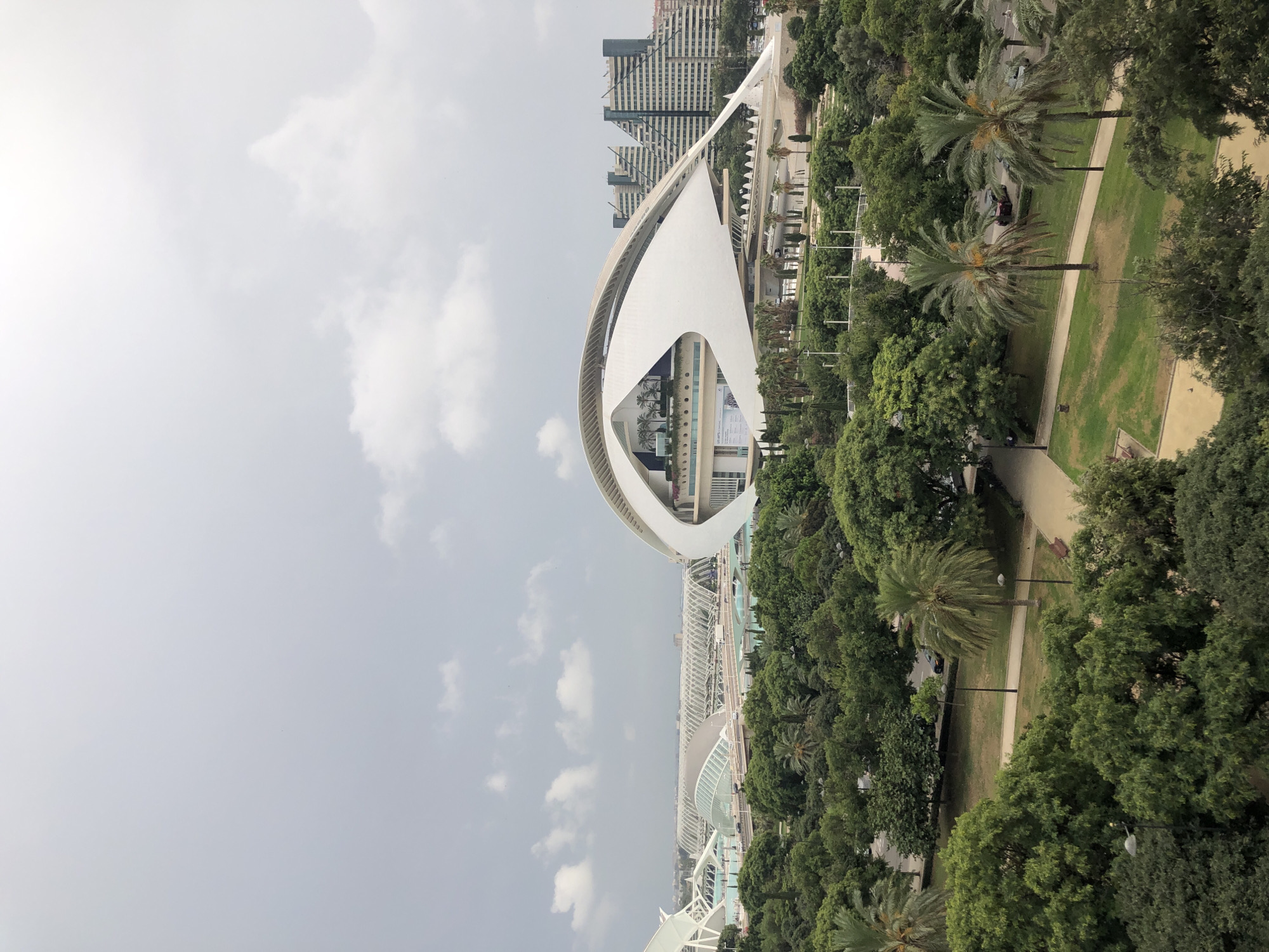 The image is an aerial view of the City of Arts and Sciences in Valencia, Spain. The foreground is dominated by lush green trees and palm trees, with pathways and park benches visible. In the mid-ground, several striking modern architectural structures are present. Most prominent is a large, white, eye-shaped building with a prominent overhang. To the left, a white, tent-like structure with translucent panels is visible. Further in the background, more buildings with geometric, stepped facades are seen, and a long, covered walkway with a curved roof stretches across the scene. The sky is overcast with scattered clouds.