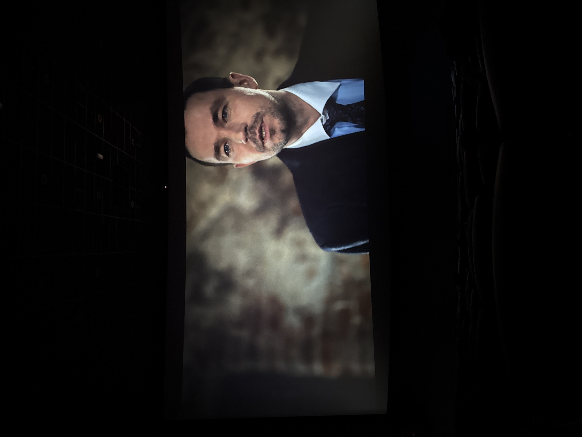A screen displaying a man in a suit and tie, likely a film or television show, is visible in a dimly lit room. The man is light-skinned with short dark hair and a trimmed beard. He wears a white collared shirt, a dark suit jacket, and a dark tie. The background behind the man is blurred. To the left of the screen, a dark surface with a grid-like pattern is present. The right side of the image is mostly dark, with hints of what appear to be people sitting in the room. The overall scene suggests a movie theater or a similar setting where a screen is being viewed in low light. There is no information that can connect the image with Perm, Russia.