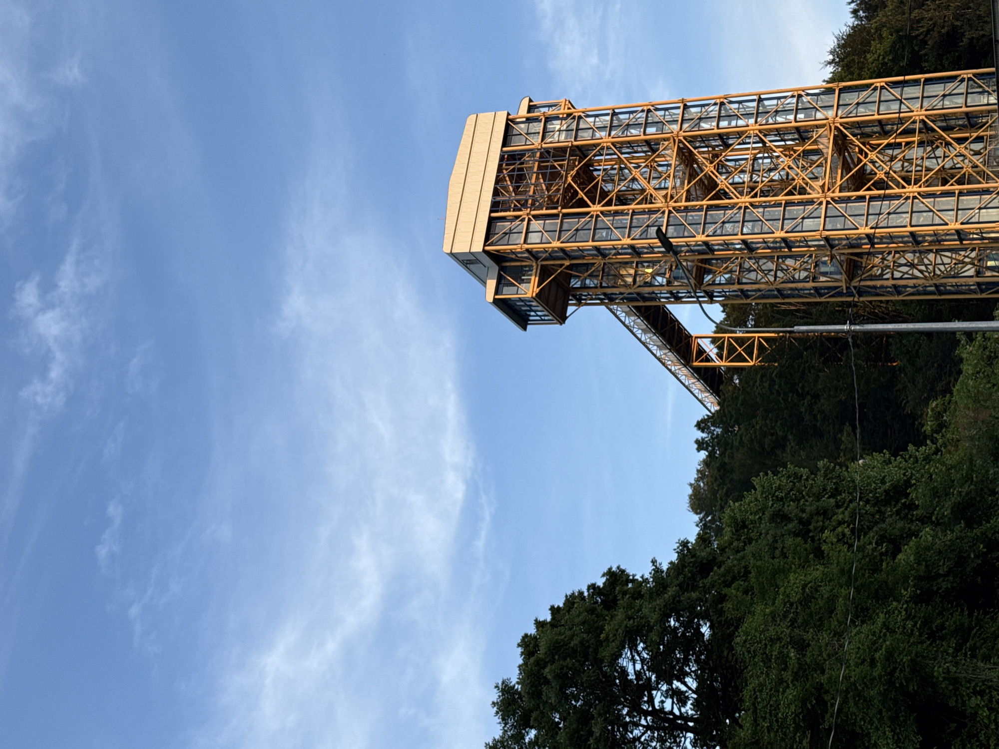 The image captures a tall, modern observation or lift tower situated amidst a densely wooded landscape in Nebug, Russia, under a clear blue sky with scattered wispy clouds. The central subject is a multi-story structure characterized by a prominent yellow-orange metal lattice framework and extensive glass paneling. The upper section of the tower features a wider, enclosed area topped with a light beige or light yellow sloped roof, suggesting a viewing platform. A pedestrian bridge or skywalk, mirroring the tower's yellow-orange structural design, extends horizontally from an elevated point on the tower towards the left, connecting it to the adjacent natural terrain. The foreground and midground on both sides of the tower are dominated by lush, dark green deciduous trees and thick foliage, indicating a forest or hilly wooded area. A utility pole with wires is partially visible on the lower left, near the edge of the tree line. Sunlight illuminates the scene, highlighting the tower's metallic structure. No individuals or specific activities are discernible within the frame.