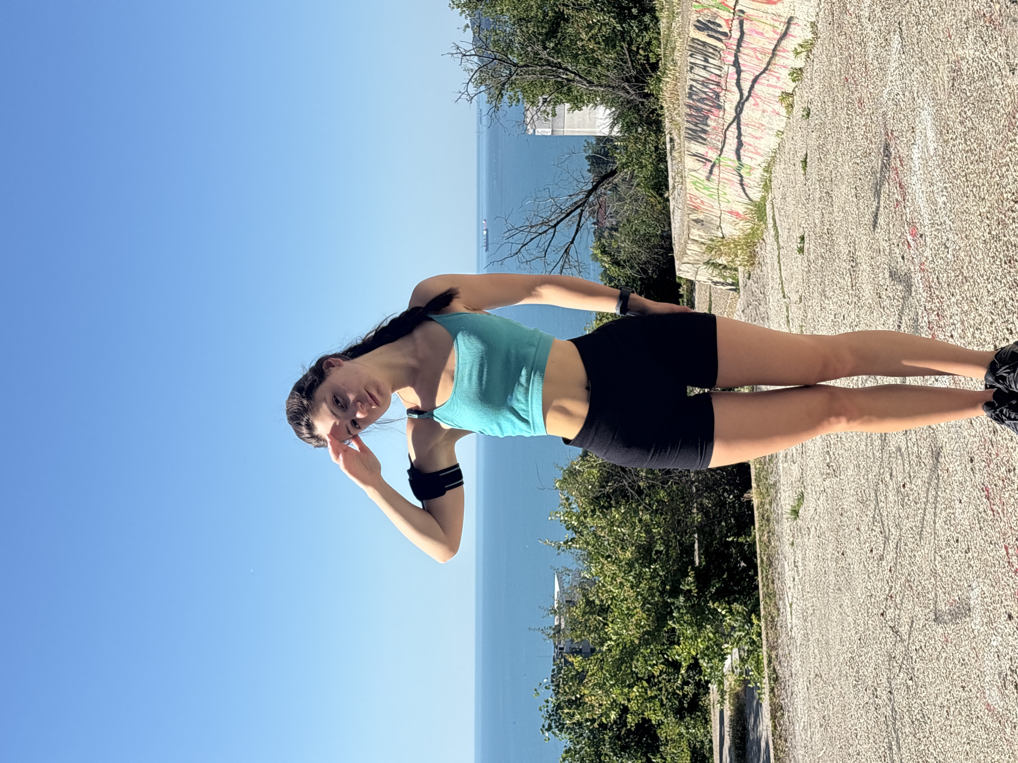 A young woman stands on a paved surface overlooking the sea. She is wearing a turquoise sports bra, black athletic shorts, and black sneakers. She has a black armband on her left arm and is shielding her eyes with her right hand, looking towards the camera. The background consists of a wide expanse of blue sea with a few ships visible in the distance. Lush green trees and some buildings are present on the coastline. The ground in the foreground is a rough, textured pavement with some areas of grass and what appears to be graffiti on a stone structure to the right. The sky is a clear, bright blue. The location is likely a vantage point in Varna, Bulgaria, offering a panoramic view of the Black Sea.