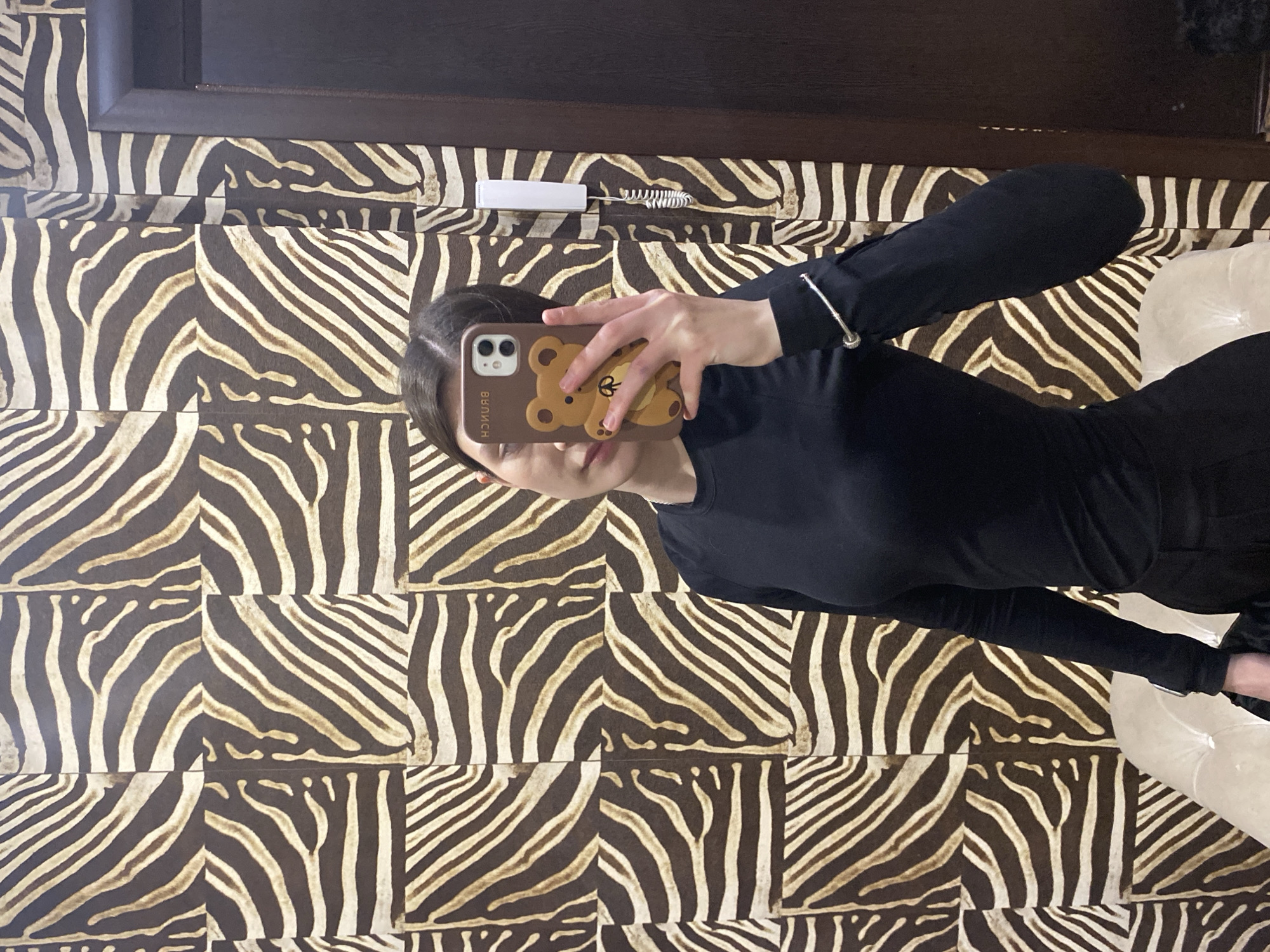 A person is taking a selfie in front of a mirror. The person is wearing a black top and black pants. The phone case is brown and has a cartoon bear on it. The wall behind the person has a zebra print pattern. A white telephone is attached to the wall. No evidence is present to confirm this is in the city of Votkinsk, Russia.