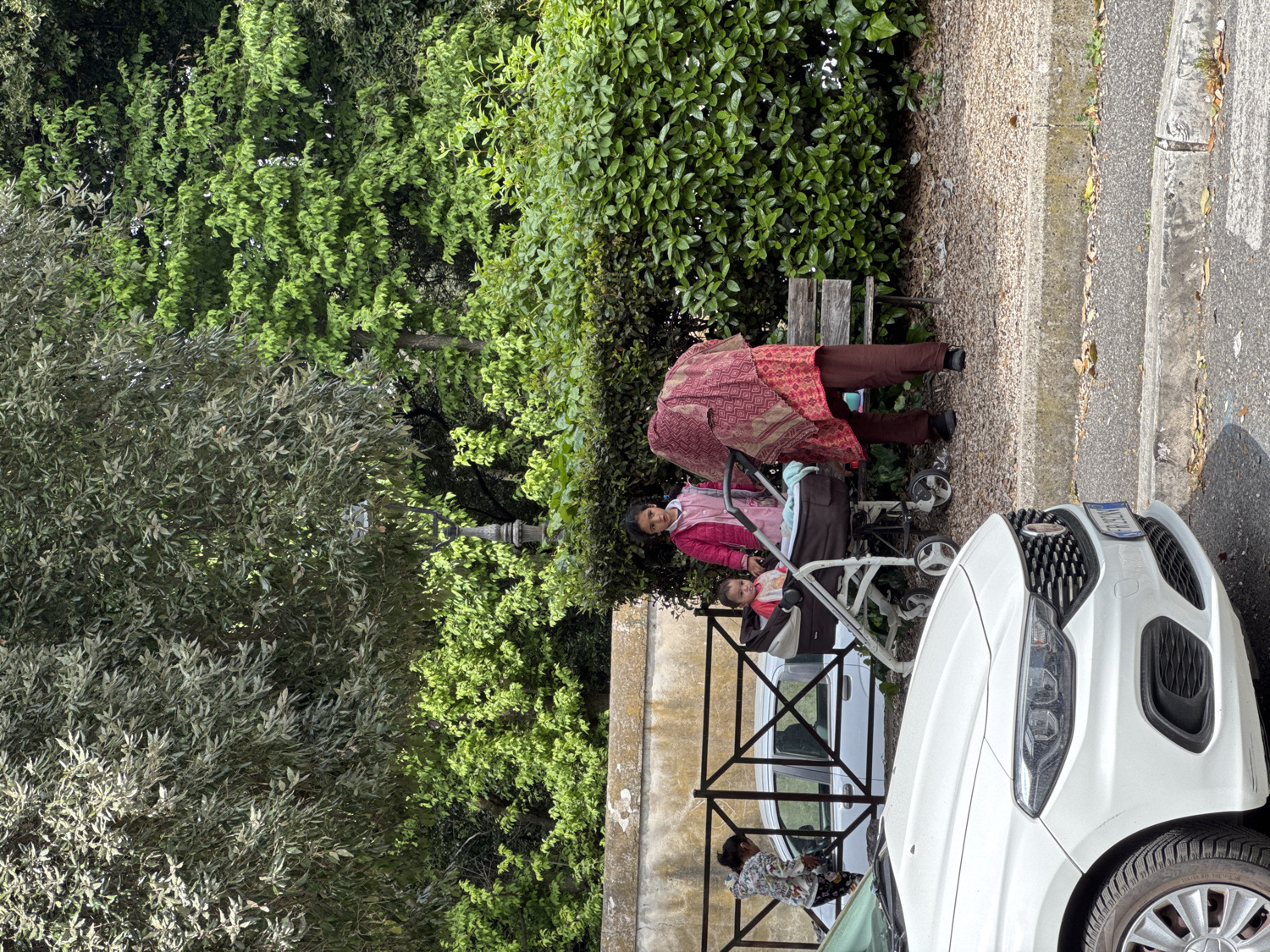 This outdoor image, captured in Ariccia, Italy, depicts a daylight scene under sunny weather conditions. The primary subjects include two women, two children, and a white car, set against a backdrop of lush greenery.
In the mid-ground, a young woman wearing a pink jacket and a light-colored top stands beside a white baby stroller. She is looking directly towards the camera. A child, wearing a red top, is seated in the stroller, also looking towards the camera. Behind them, partially obscured by dense green bushes, another woman, dressed in a red patterned top and brown trousers, is bent over. Her face is not visible. Behind a black metal fence in the foreground, partially obscured by the white car, another child wearing a patterned top can be seen.
A white car is parked on an asphalt road, with its front and passenger side visible on the right. Its Italian license plate clearly shows "EA 749 EH". A concrete curb separates the road from a paved pathway and a gravel strip leading to a continuous hedge of green bushes. A wooden bench is visible within the bushes. The background is dominated by a variety of trees and thick foliage in different shades of green, with a street light pole visible among them. The bright sunlight creates clear shadows, indicating it is either morning or afternoon. The overall impression is one of a quiet, casual gathering alongside a road or park path.