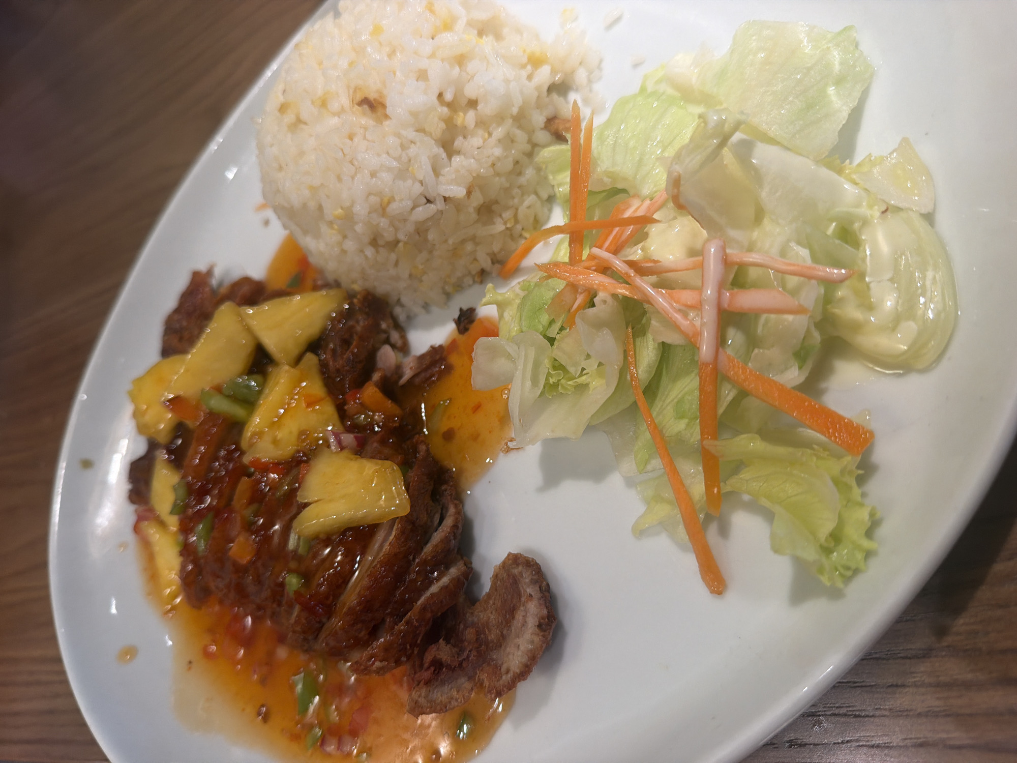 A plate of food is presented on a wooden table. The plate contains sliced duck covered in a sweet and sour sauce with pineapple chunks and what appear to be small pieces of red pepper and green onions. To the right of the duck is a mound of white rice. In the lower right corner, a small salad of lettuce and shredded carrots with a creamy dressing is visible. The food is served on a white oval plate. The lighting suggests it may be indoors, possibly a restaurant. The location is unknown.