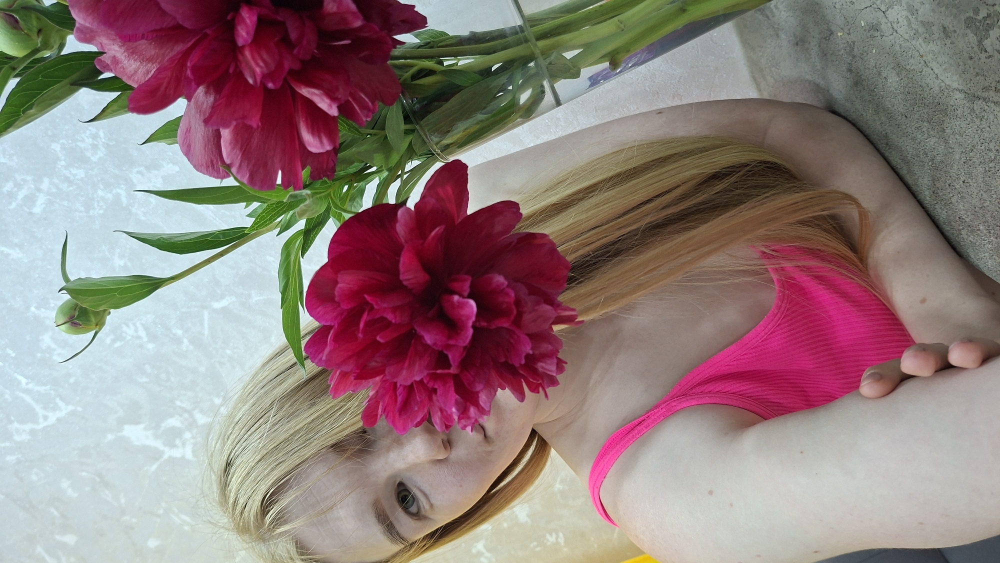 A medium shot captures a fair-skinned person with long, straight blonde hair and bangs, wearing a vibrant neon pink ribbed tank top. The left side of the person's face is visible, showing a light-colored eye and some freckles, with the mouth area partially obscured by a large, open, dark pink peony bloom positioned directly in front of the face. To the right of the person, a clear glass vase holds several stems of dark pink peonies, including more open blooms and multiple green buds with green leaves. The person's arm rests on a light grey, textured surface in the foreground. The background features a light-colored wall with a subtle, textured, patterned design. The scene is indoors.