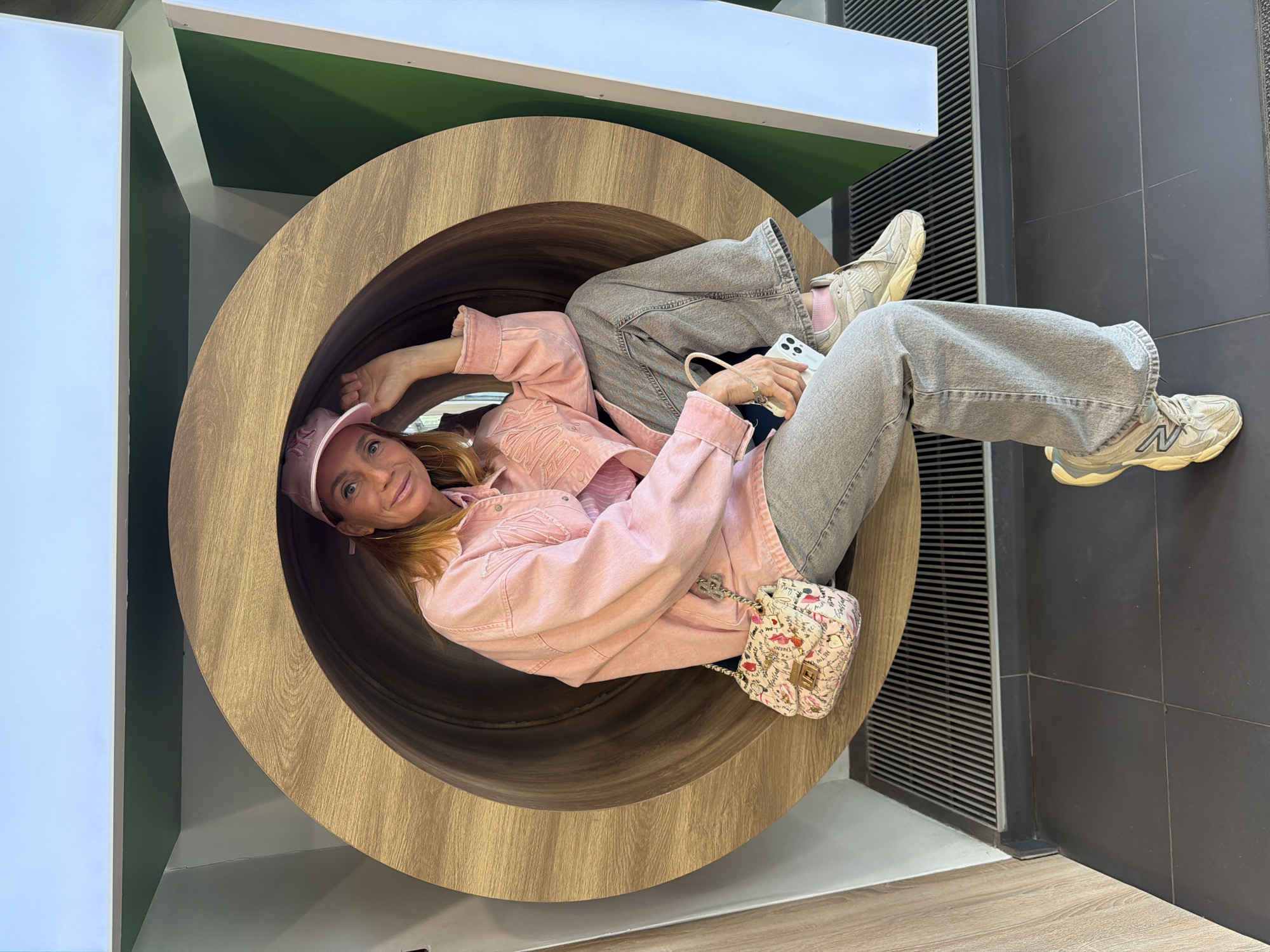 A blonde woman wearing a pink baseball cap, pink denim jacket, light gray jeans, and light gray New Balance sneakers, is reclining inside a large wooden cylinder. She has a small, pink, floral-patterned purse slung across her torso and is holding a white smartphone. The cylinder appears to be a design element in an interior space. The background includes sections of white, green, and gray walls, as well as a section of dark gray tiled floor with a vent. The scene is indoors and well-lit. The location is identified as Saint Petersburg, Russia.