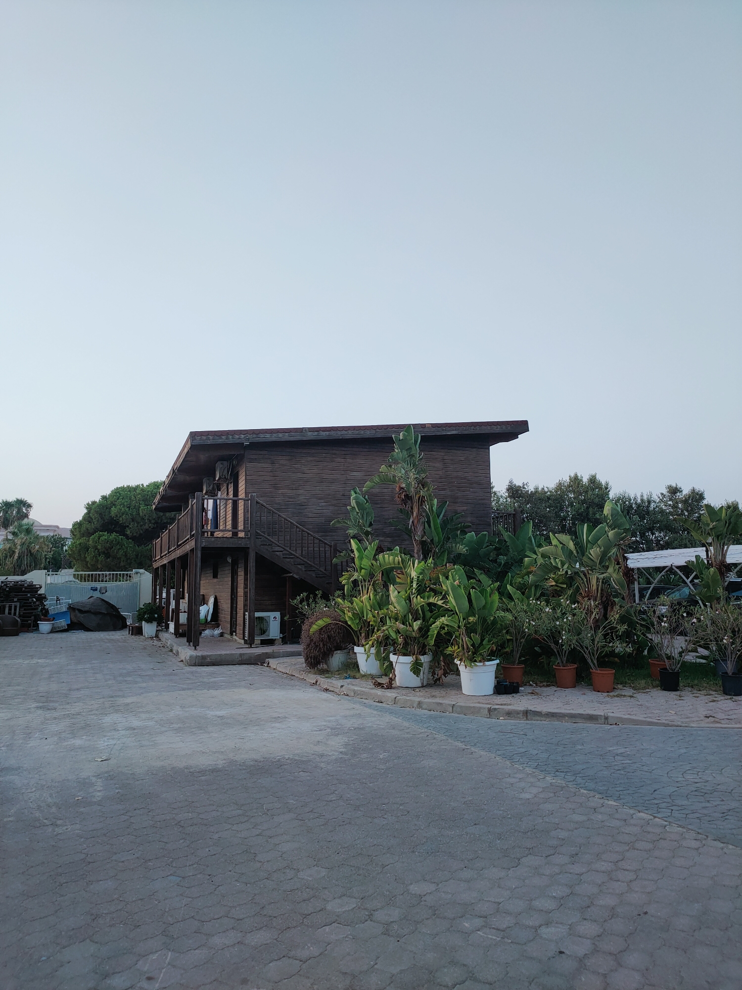 A wooden building with an elevated porch is situated in an outdoor paved area. The building has a pitched roof and a dark wooden exterior. A staircase leads up to the porch, which is lined with a wooden railing. Several air conditioning units are visible on the exterior walls of the building. In front of the building, there is a collection of potted plants of various sizes, with lush green foliage. The ground is paved with interlocking concrete stones, and a curb separates the paved area from a patch of grass where more potted plants are located. In the background, there are trees and what appears to be a fence. The sky is clear and pale, suggesting it is either early morning or late afternoon. The scene appears to be in a commercial or residential area, possibly a cafe or guesthouse, with a focus on outdoor greenery. The location is specified as Nabeul, Tunisia.